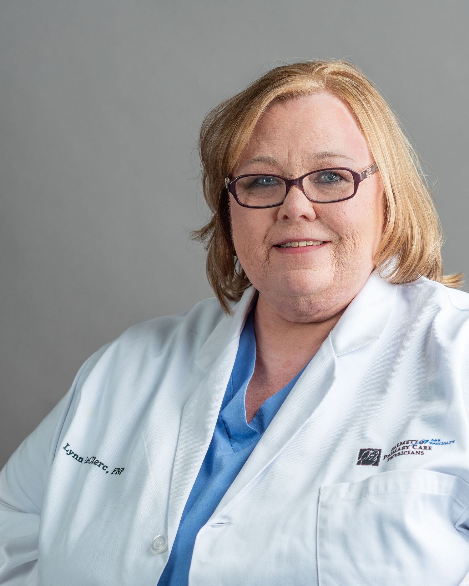 Female healthcare professional in white lab coat and blue scrubs with glasses, smiling in a professional portrait photo.