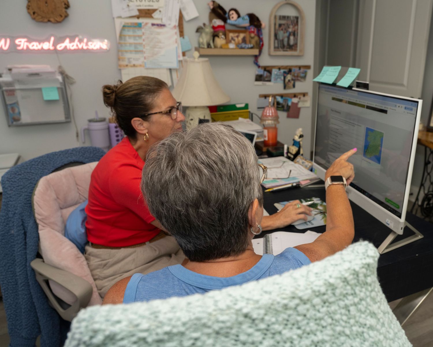 Two women collaborate at a desktop computer in a home office, one pointing at the screen while reviewing travel plans.