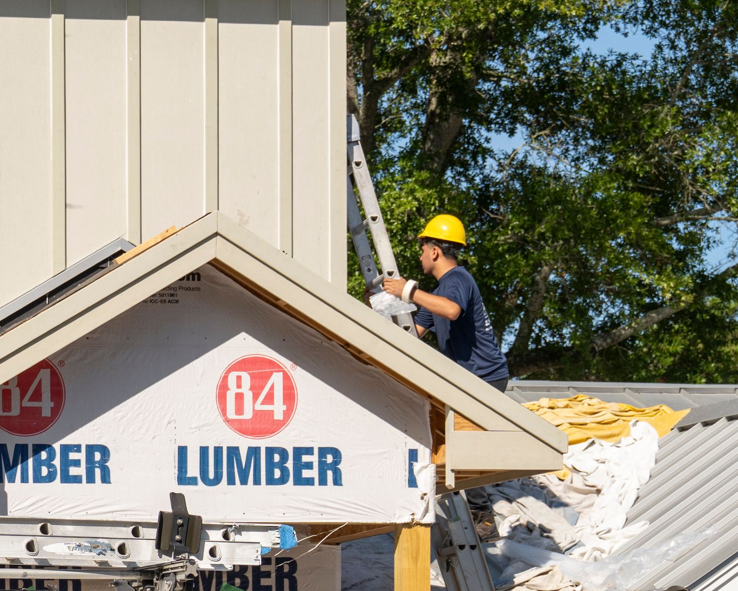 Worker in yellow hard hat installs roofing on a building framed with 84 Lumber materials at a construction site.