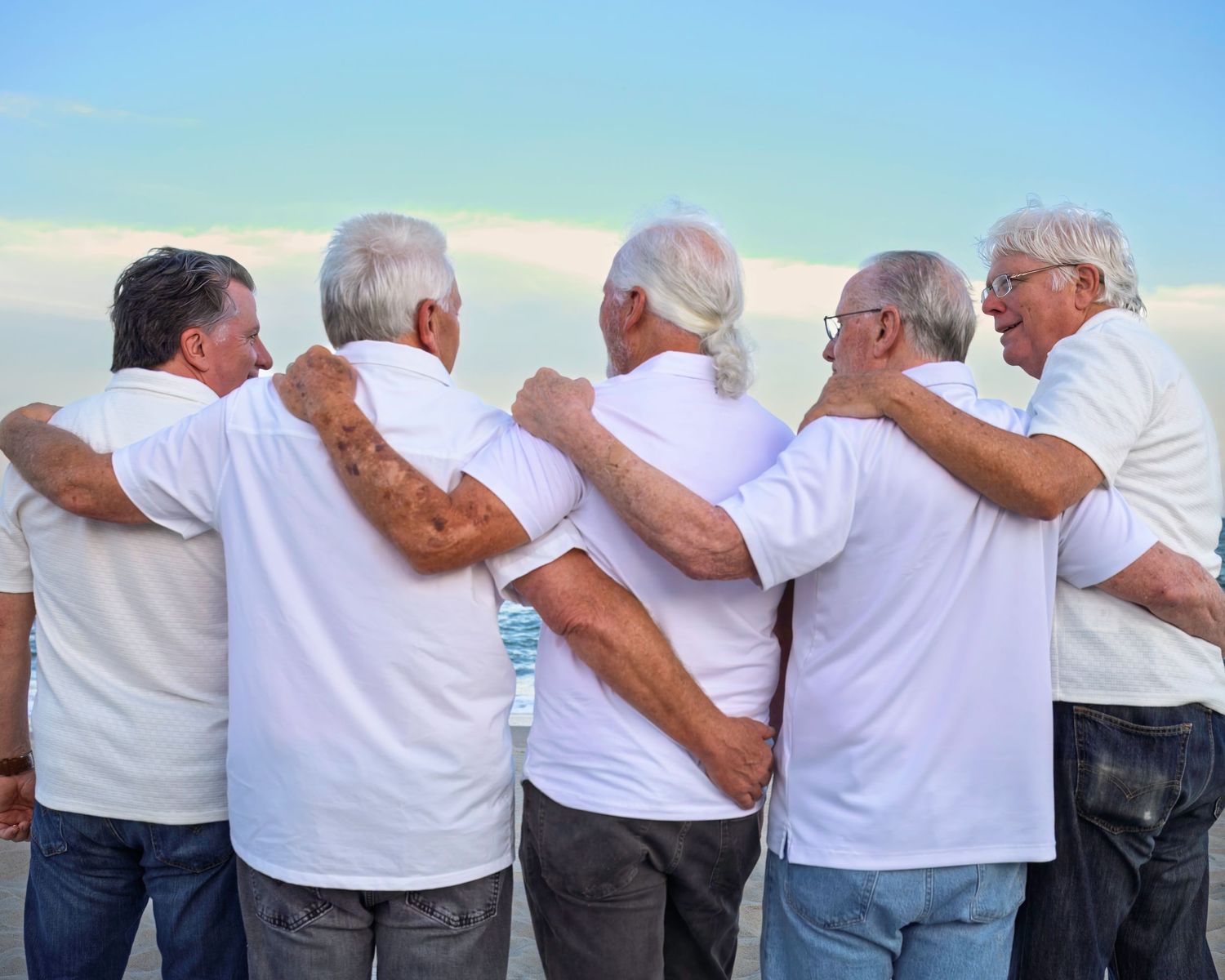 A group of people in white shirts and gray pants stand with their arms around each other looking at the ocean.