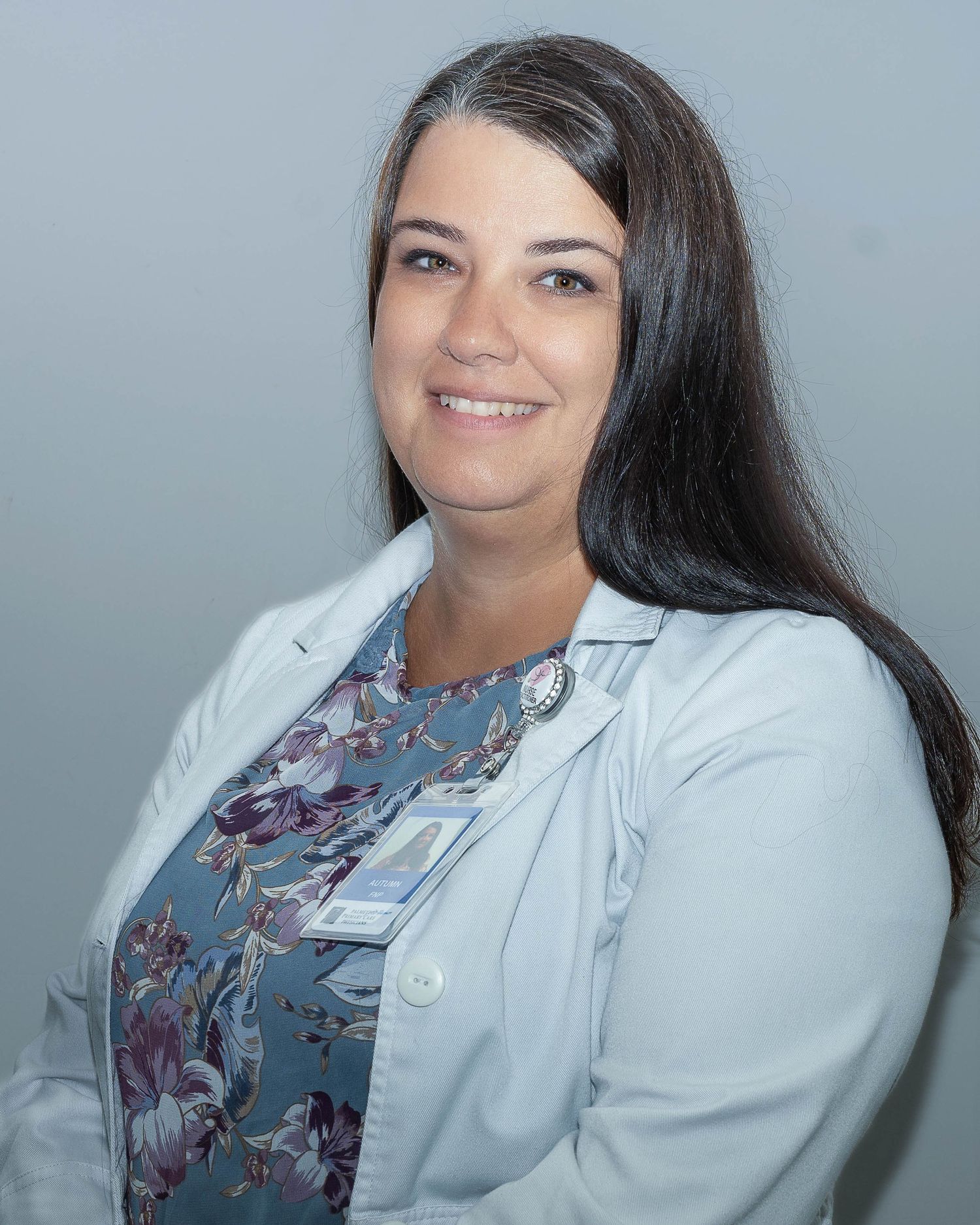 Female healthcare professional in white lab coat and floral scrubs smiling confidently against gray background.