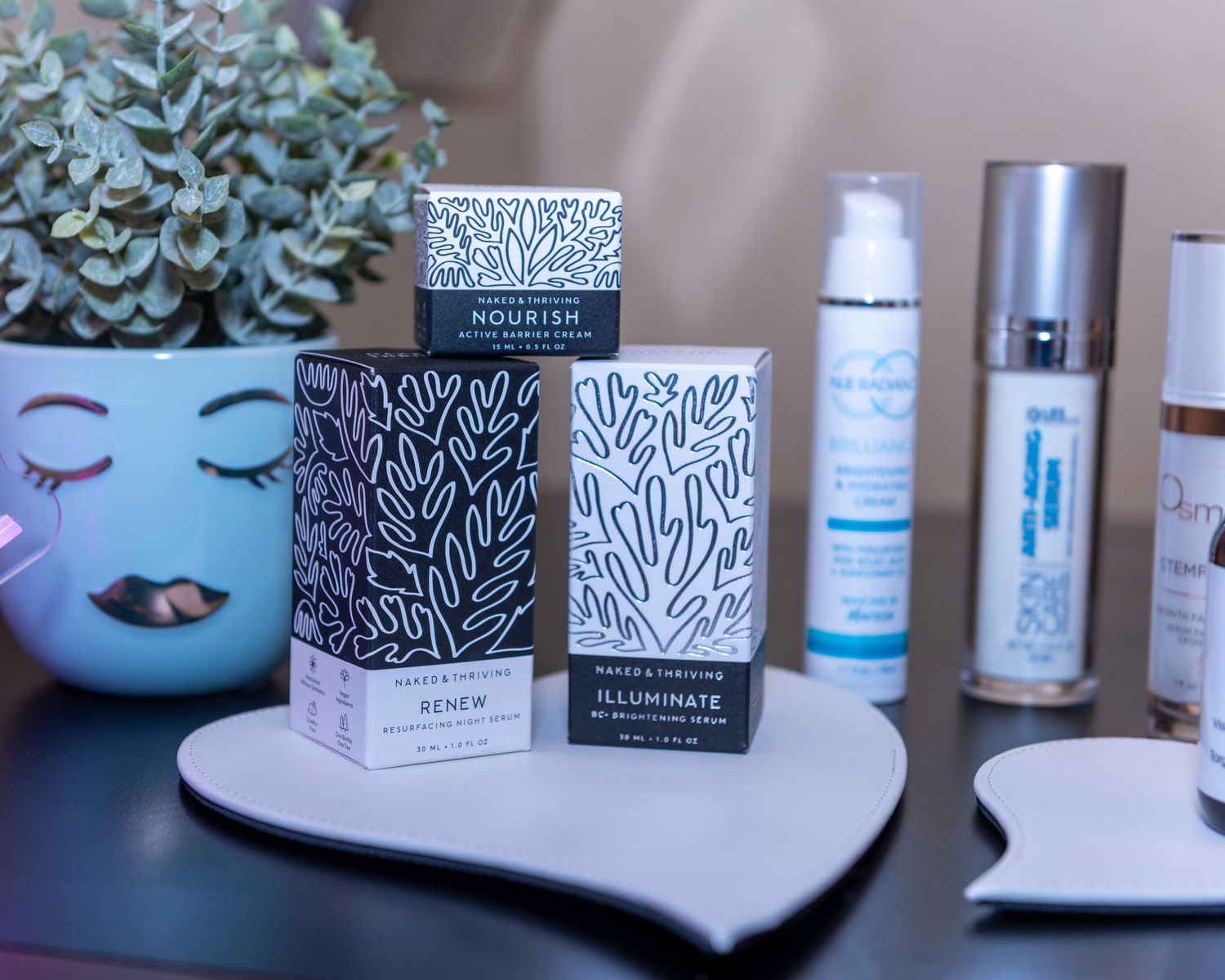 Skincare products including Nourish and Illuminate serums displayed on a black tray beside a decorative plant.
