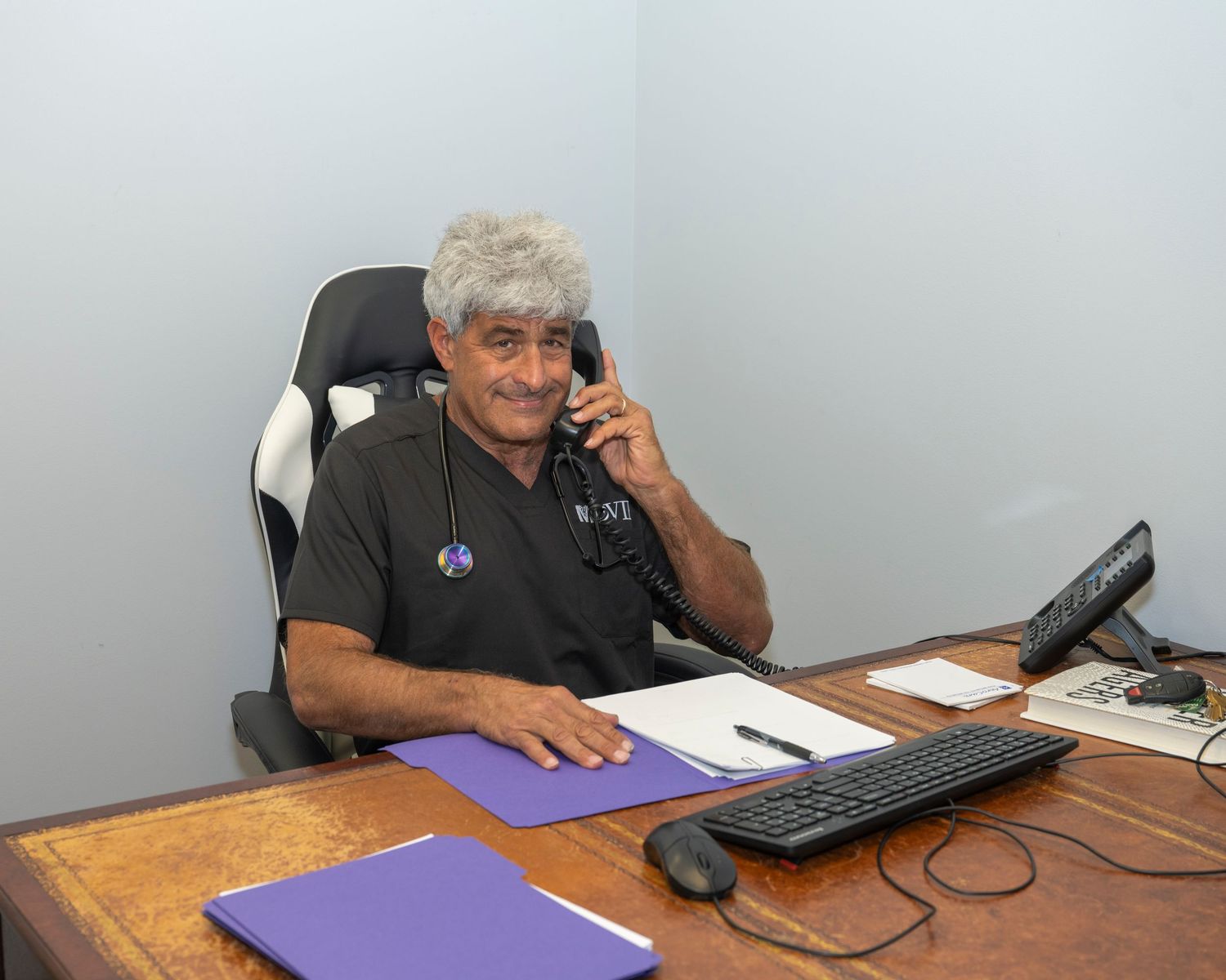 Smiling man sitting at office desk talking on phone with keyboard and purple folder nearby.