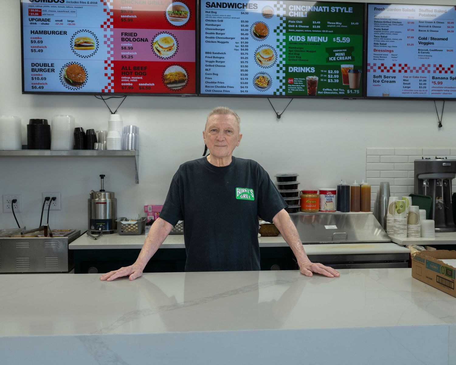 Man in black t-shirt stands behind counter of fast food restaurant with menu boards displaying sandwiches and drinks.