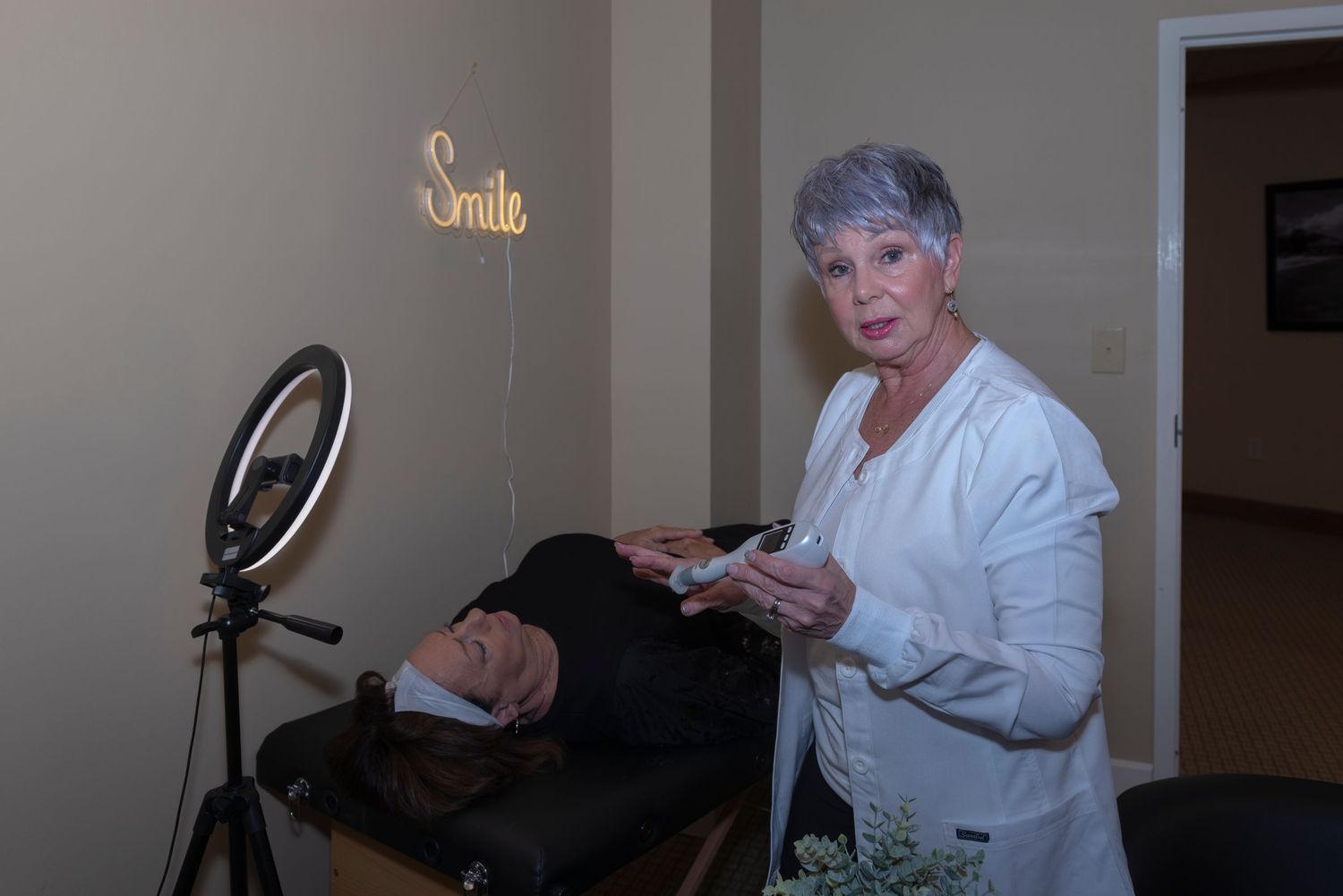 Esthetician performing a facial treatment on a client in a beauty salon with a ring light and neon Smile sign.