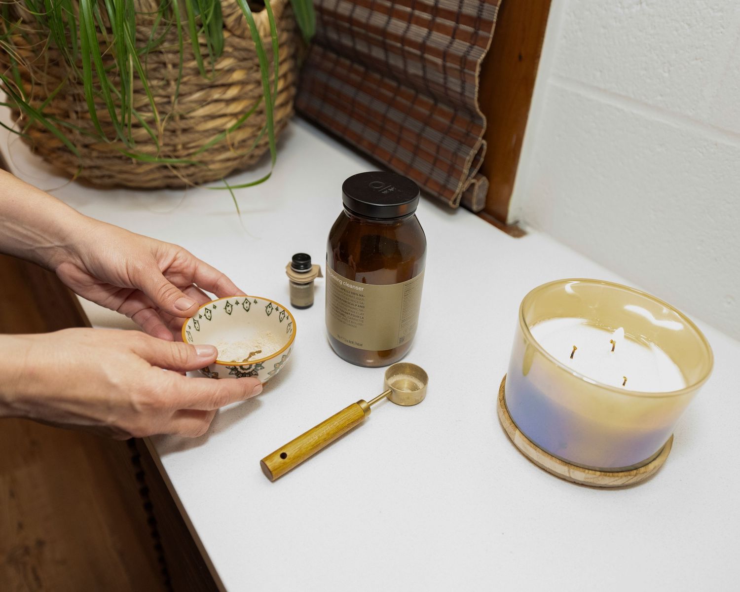 Hands holding a ceramic bowl near a candle, amber bottle, and gold spoon on a white surface.