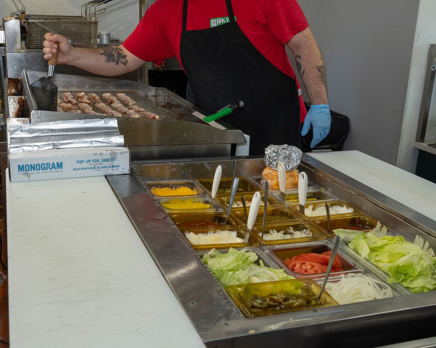 A cook prepares burgers at a fast food counter with condiment trays of lettuce, tomatoes, and sauces.
