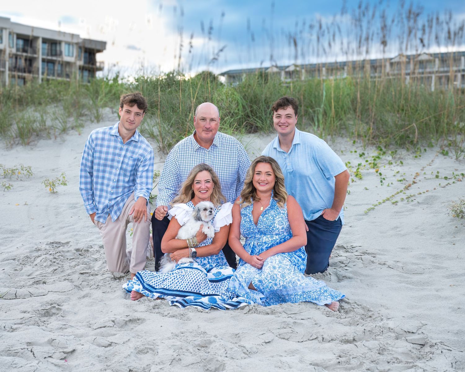 A group wearing coordinated blue and white outfits poses together on a sandy beach with sea grass in the background.