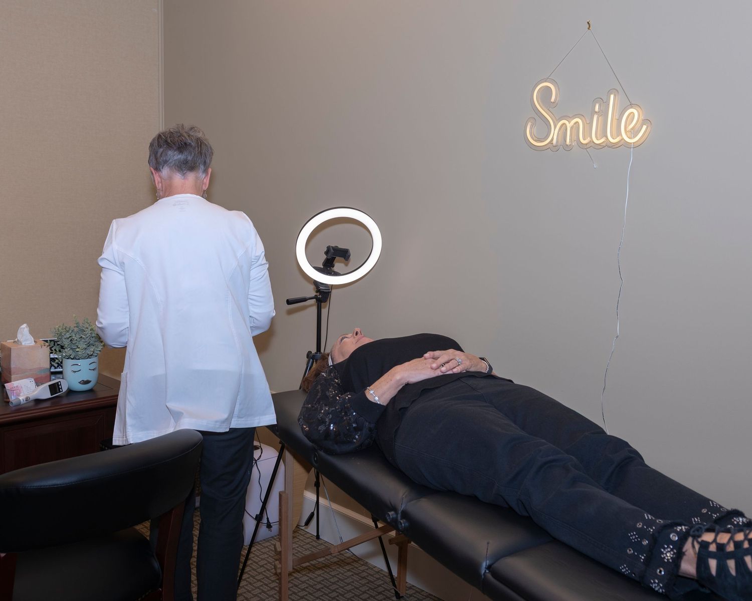 Medical professional in white coat prepares treatment area with ring light beside patient lying on table under Smile sign.