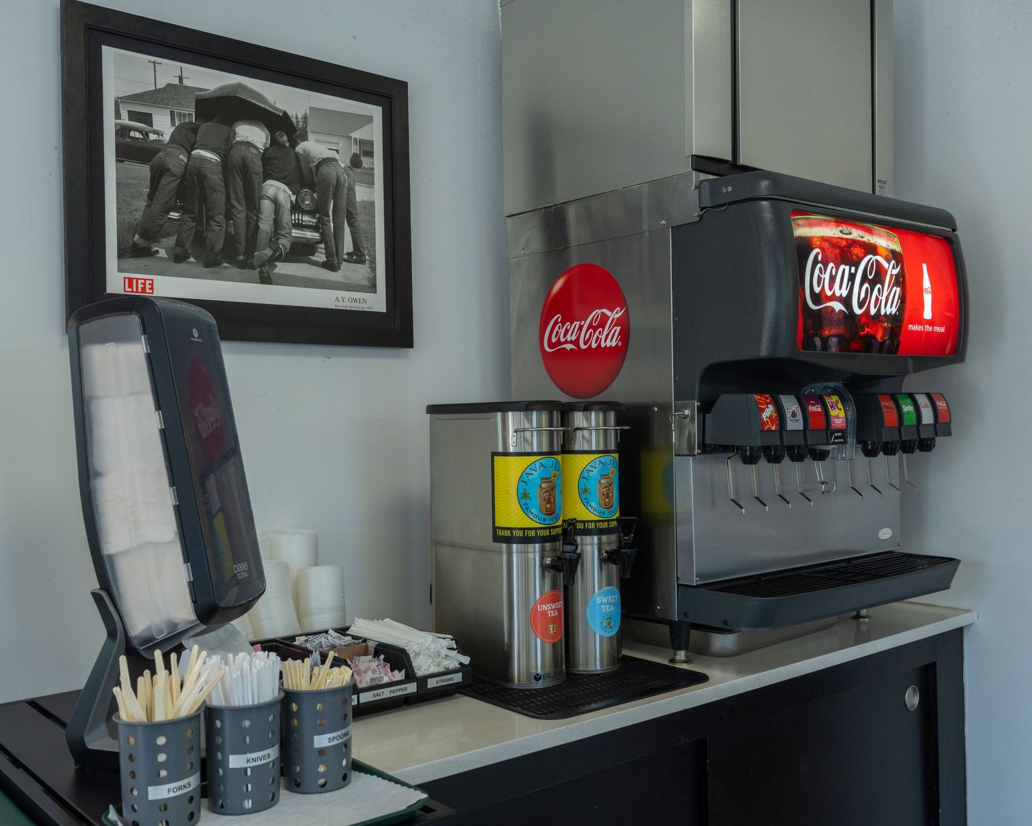 Coca-Cola fountain drink dispenser with cup holders and stirrers on a counter beside a vintage black and white photo.