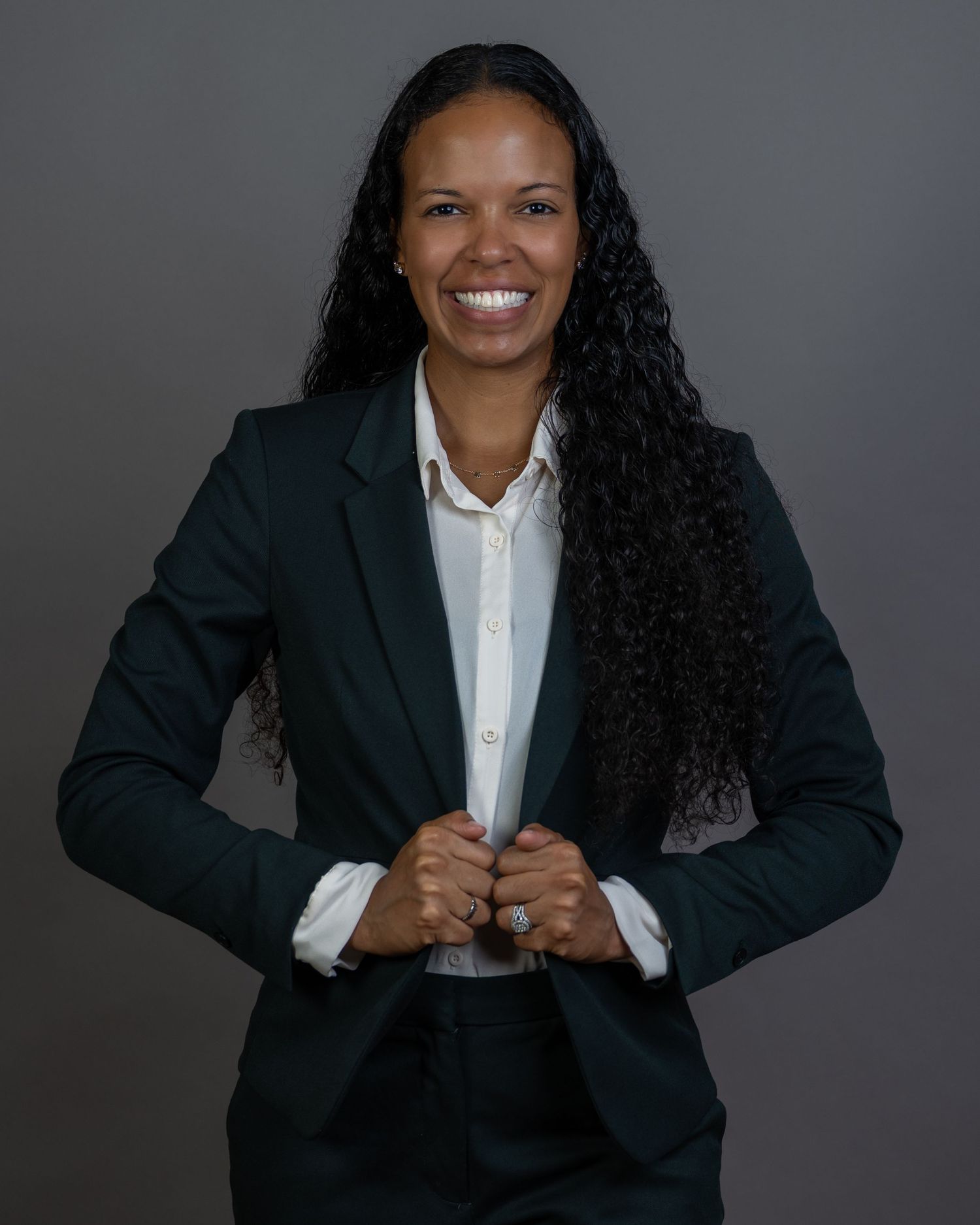 Professional woman in a dark green blazer with long curly hair smiling confidently against a gray background.