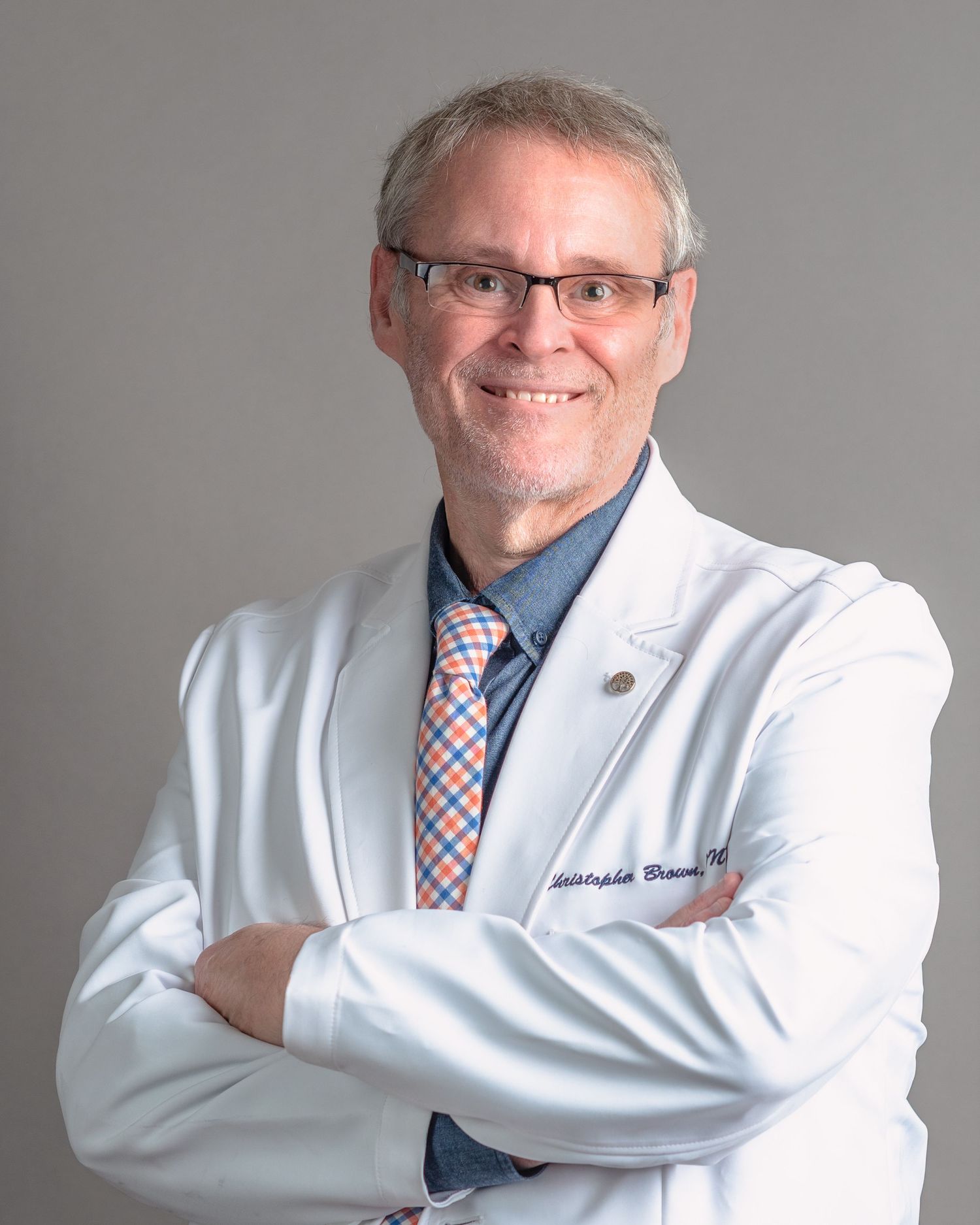 Smiling male doctor in white lab coat with arms crossed, wearing a plaid tie, professional medical portrait.