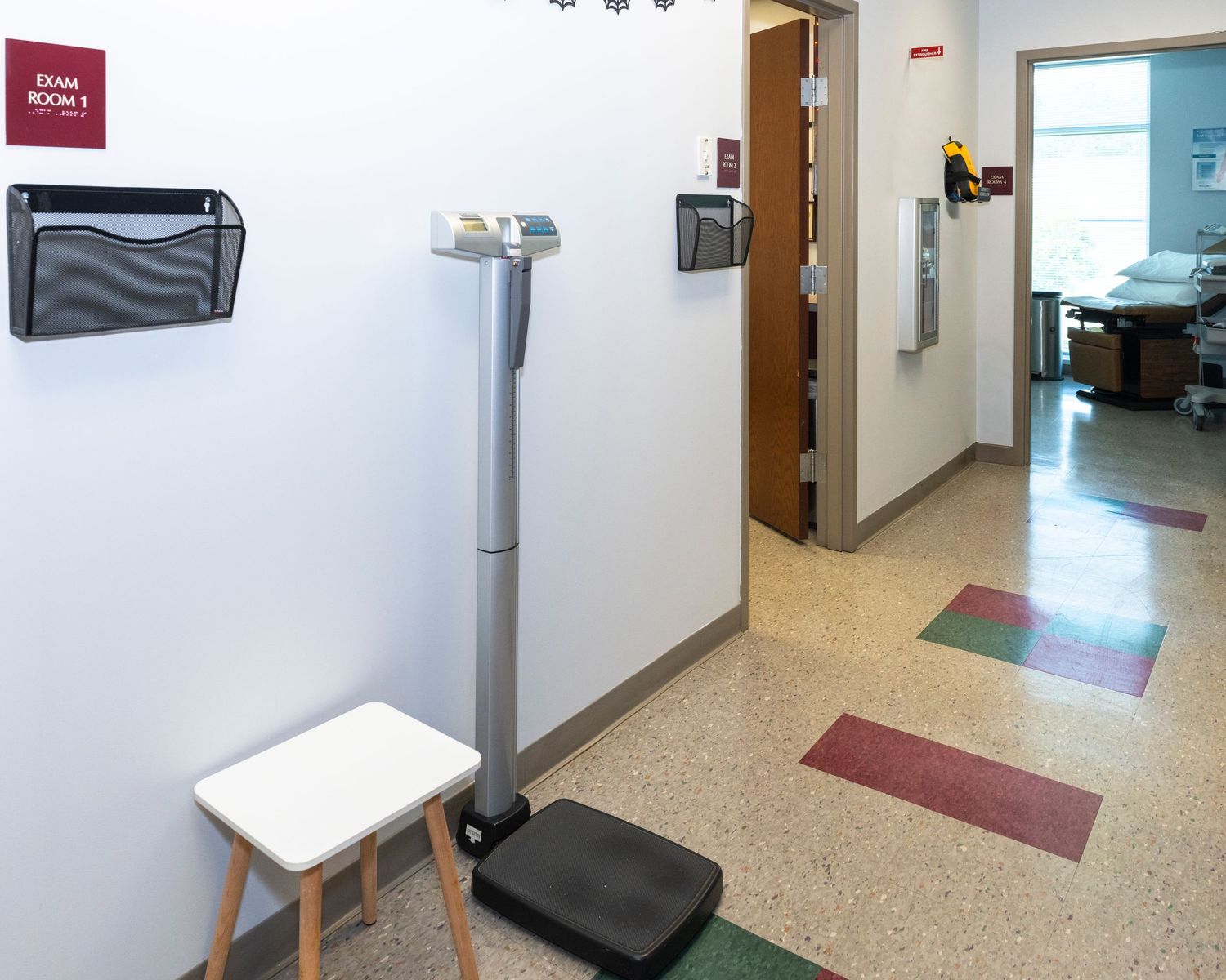 Medical exam room hallway with scale, step stool, and colorful floor tiles in a clinical healthcare facility.