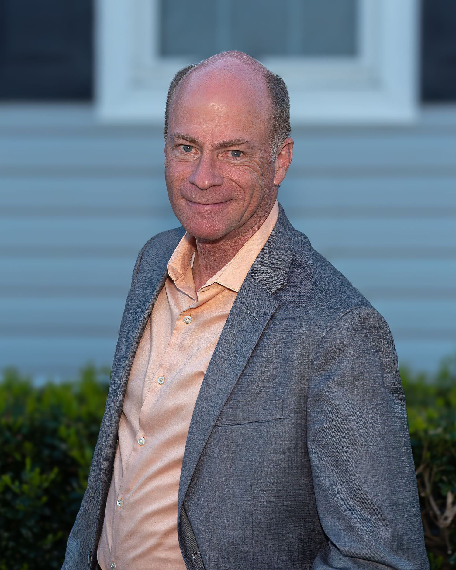 Professional headshot of a smiling middle-aged man in a gray blazer and light pink shirt outdoors.