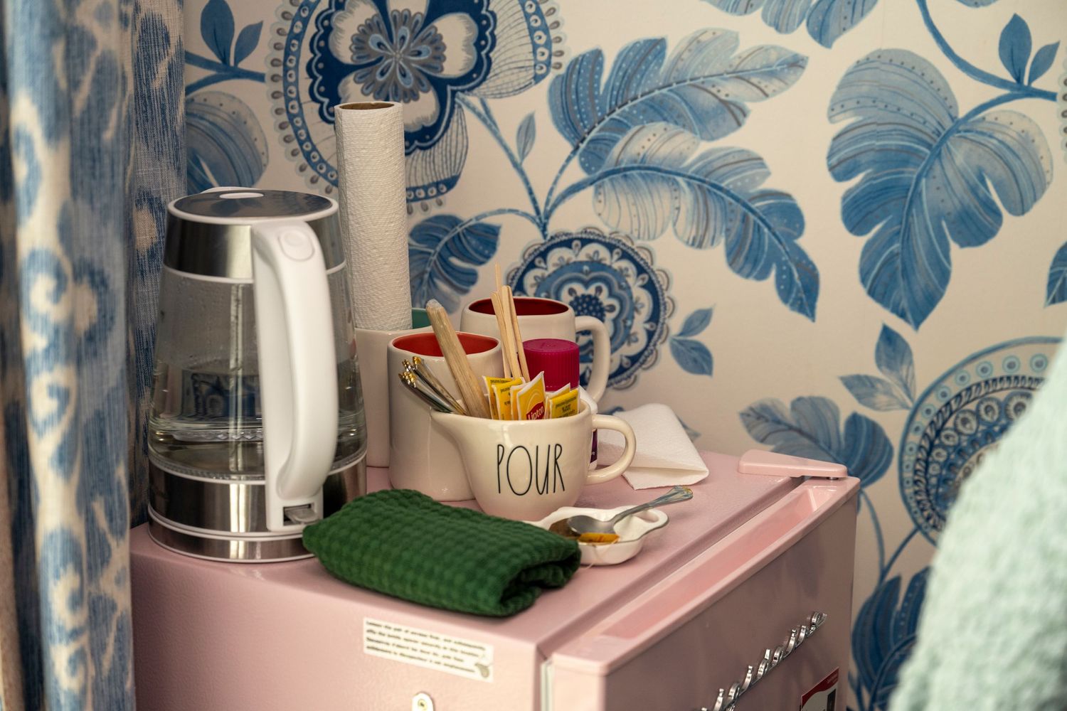 Pink mini fridge with electric kettle, floral wallpaper, and a mug holder with utensils in a cozy kitchen corner.
