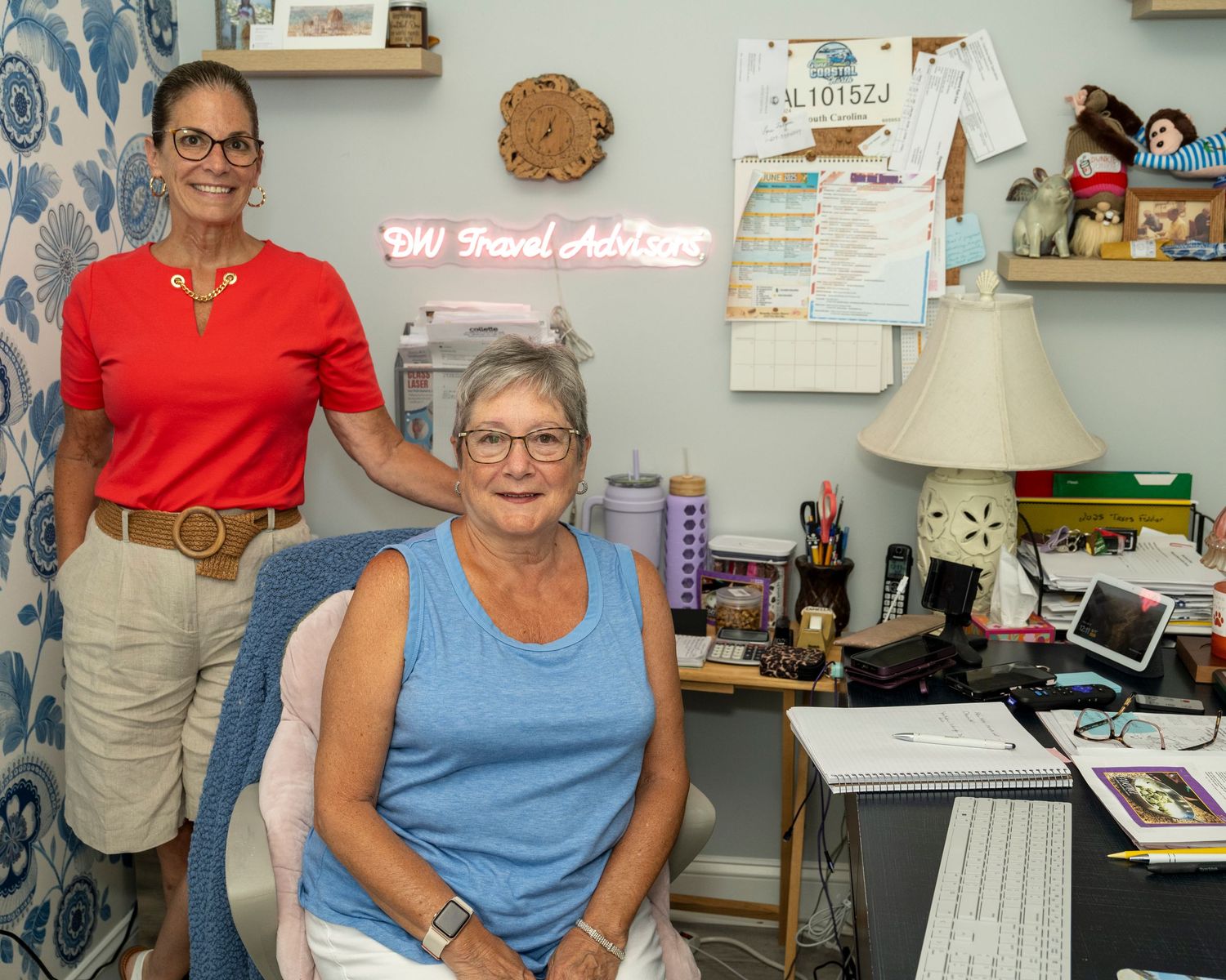 Two women smile in a cozy home office with a desk, laptop, lamp, and a BW Travel Advisor neon sign on the wall.