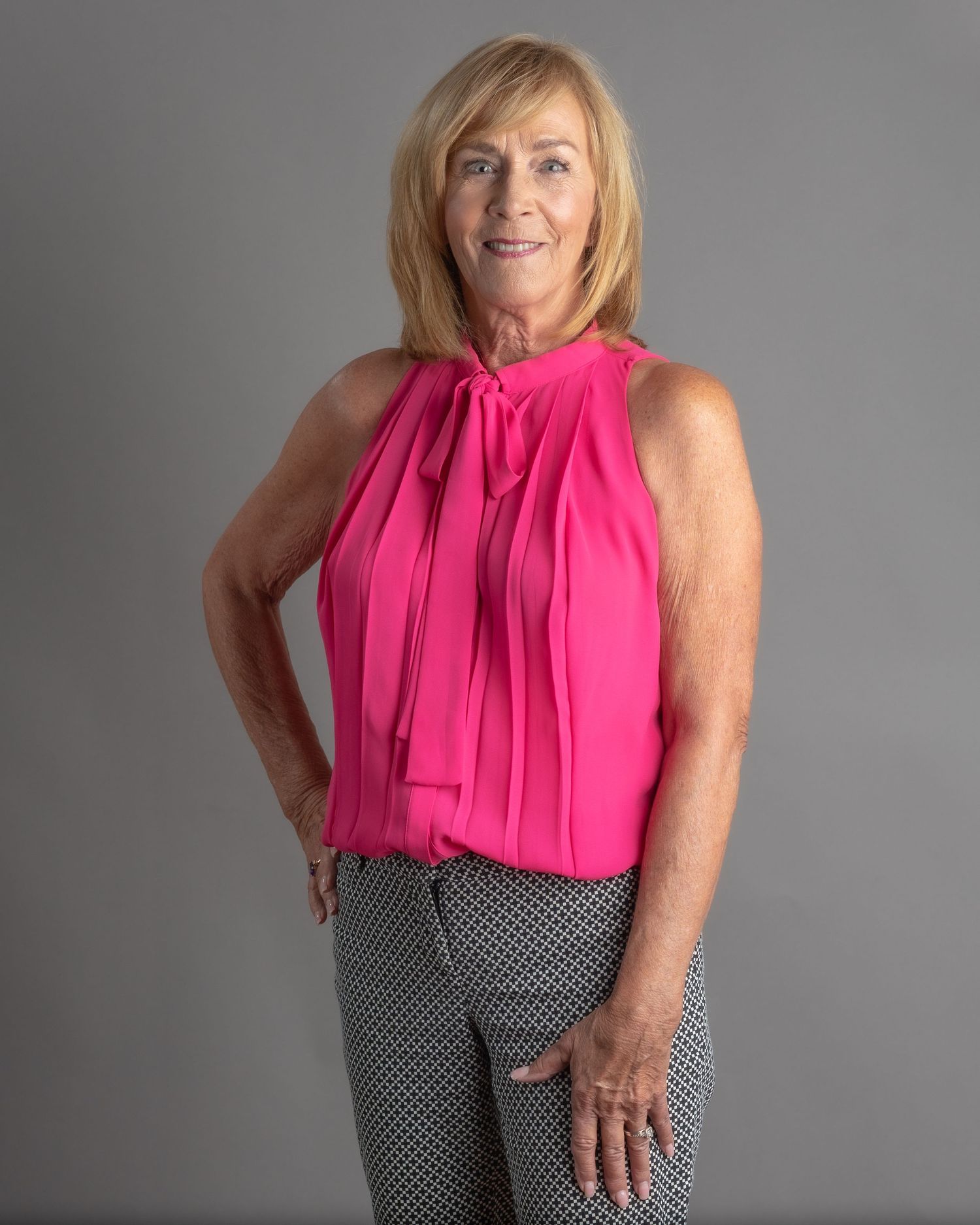 Woman in pink sleeveless top and patterned pants posing confidently against a gray studio background.