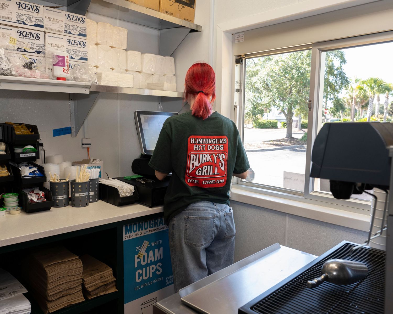 Red-haired worker in Bubba's Grill shirt stands at counter inside small restaurant kitchen near window.