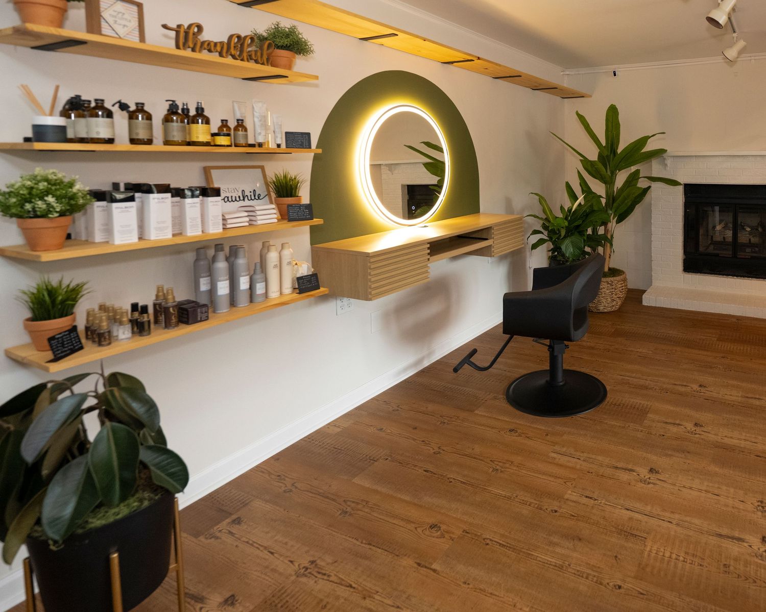 Modern hair salon interior with illuminated round mirror, wooden shelves with products, styling chair, and lush green plants.