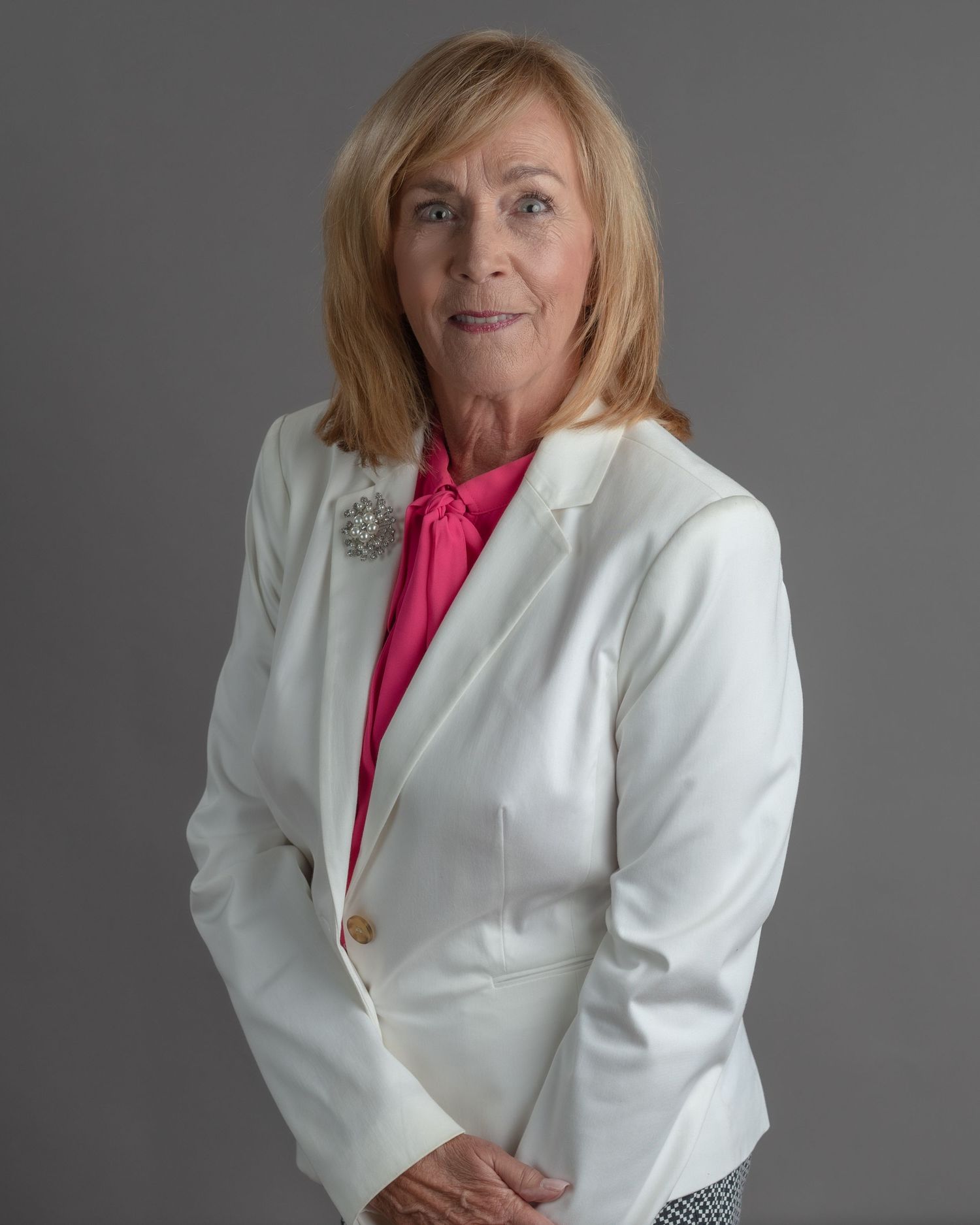 Professional woman in white blazer and pink blouse with brooch poses for formal portrait photo.