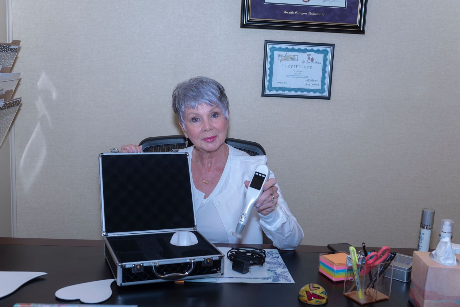 A woman with gray hair sits at a desk holding a device next to an open case with medical certificates on the wall.