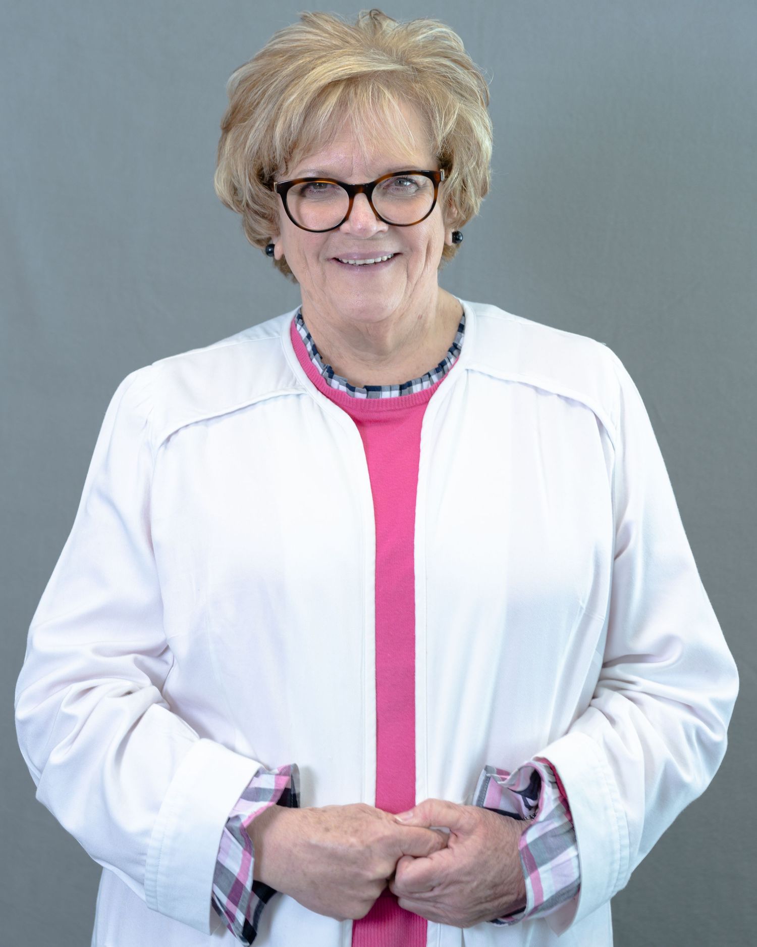 Smiling woman in white lab coat and pink top with glasses, posing professionally against gray background.