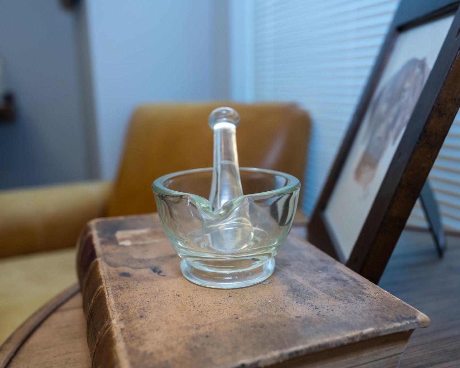 Clear glass mortar and pestle set resting on an antique book, with a leather chair and framed art in the background.