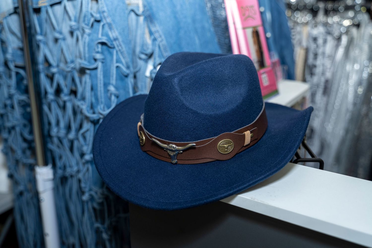 Navy blue cowboy hat with brown leather band displayed on a shelf in a western accessories store.
