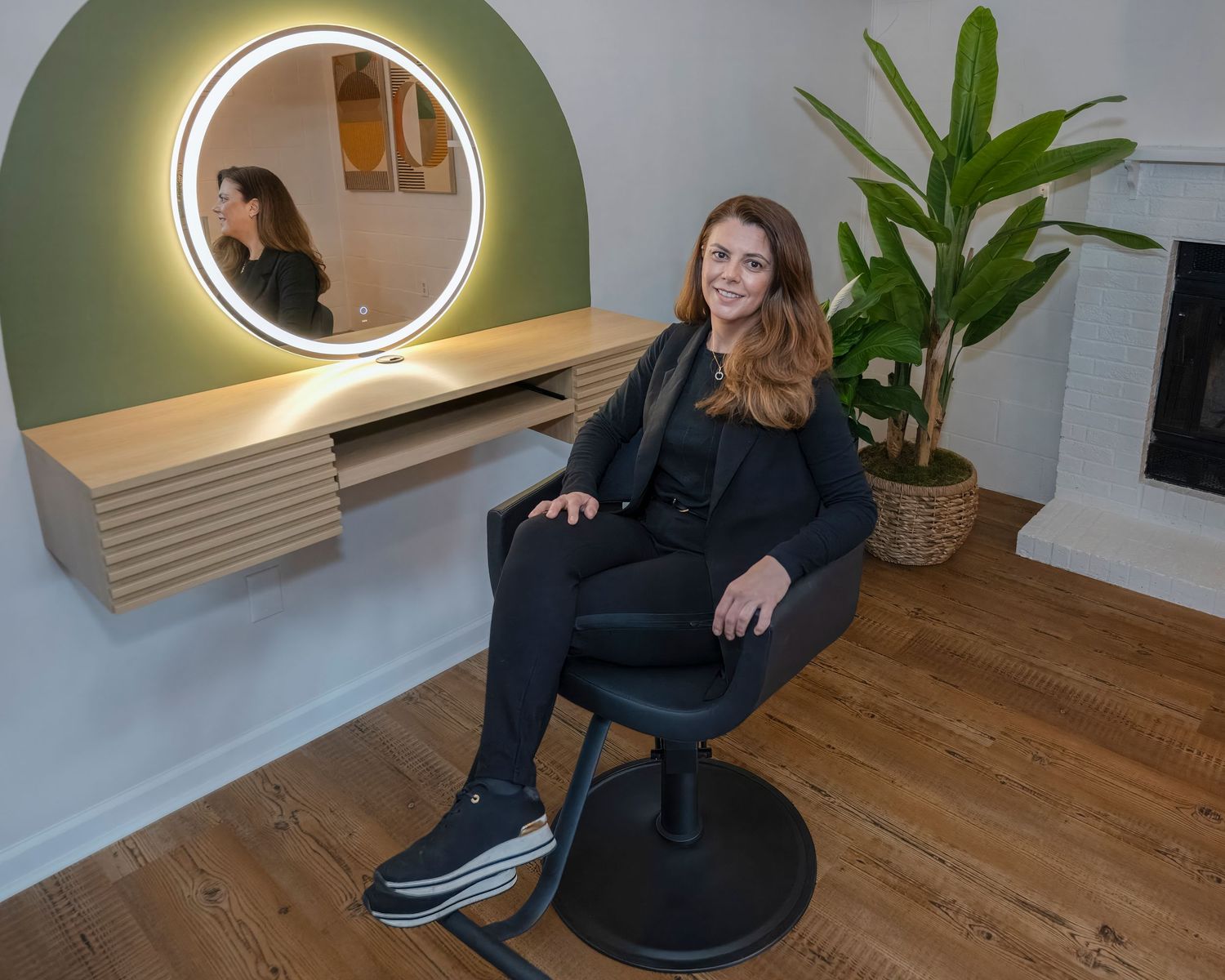 Woman sitting in salon chair beside illuminated round mirror and green accent wall in modern hair salon.