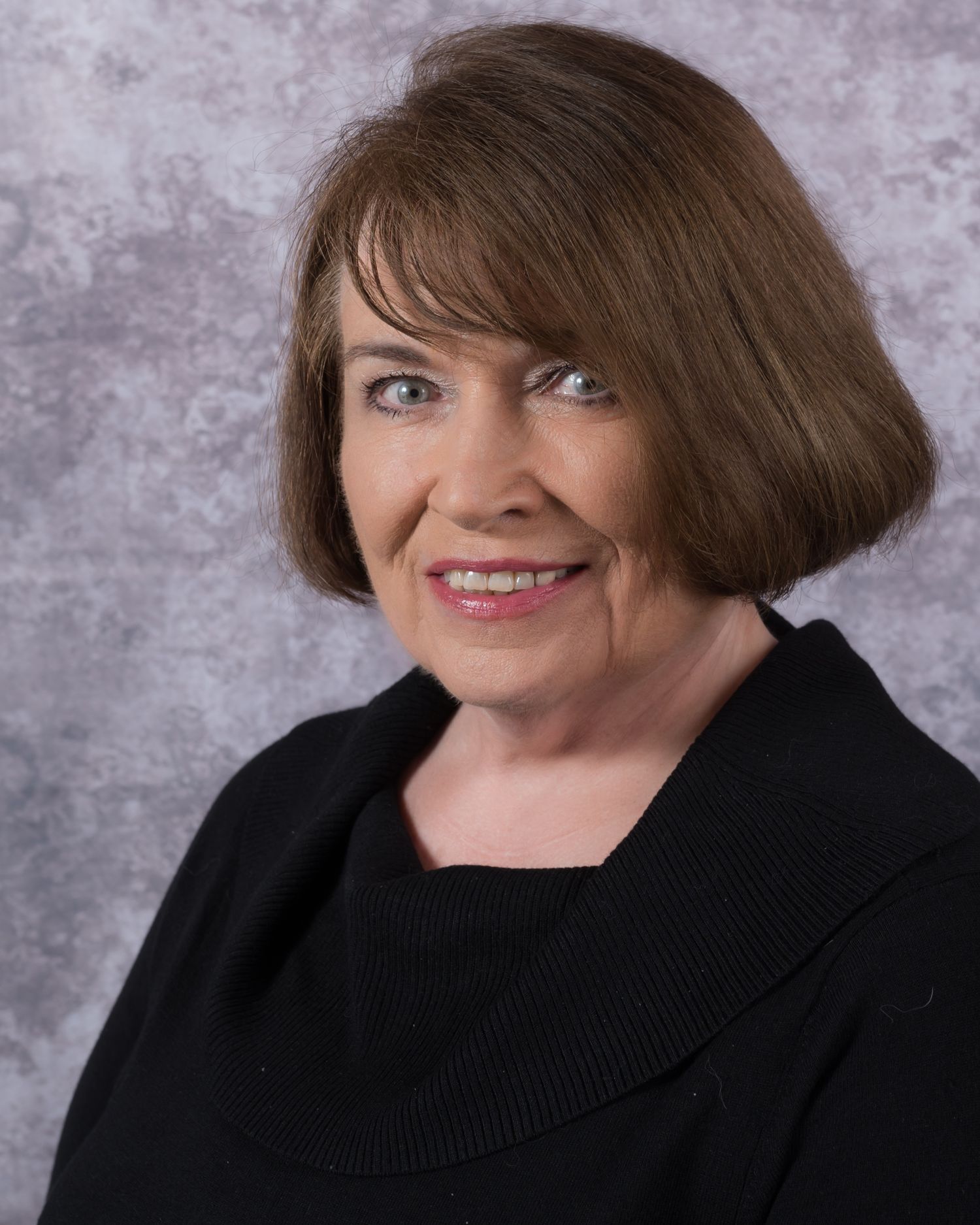 Professional headshot of a smiling middle-aged woman with brown bob haircut wearing a black cowl-neck top.