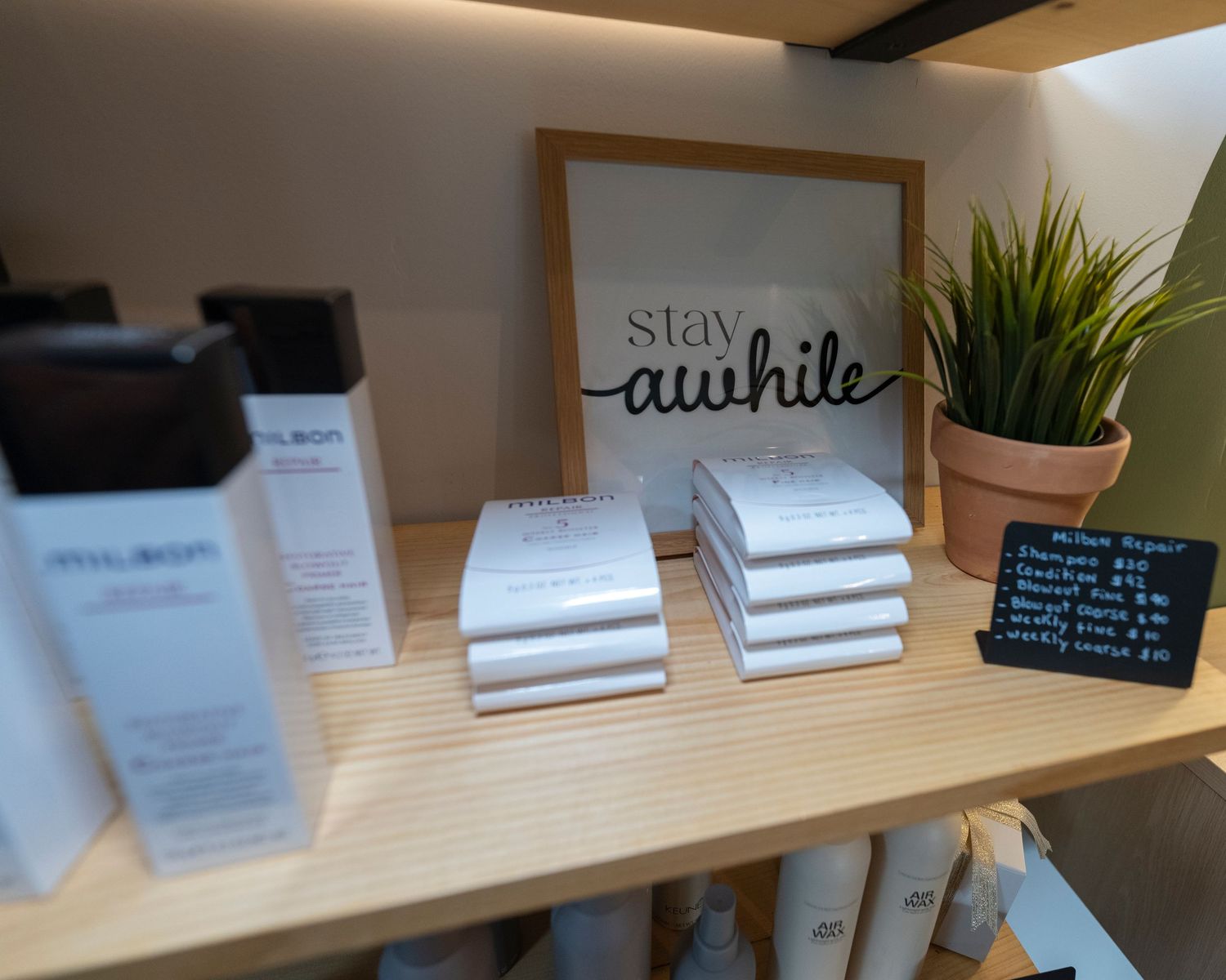 Wooden shelf display with skincare products, stacked notepads, a 'Stay Awhile' sign, and a green plant in a salon.