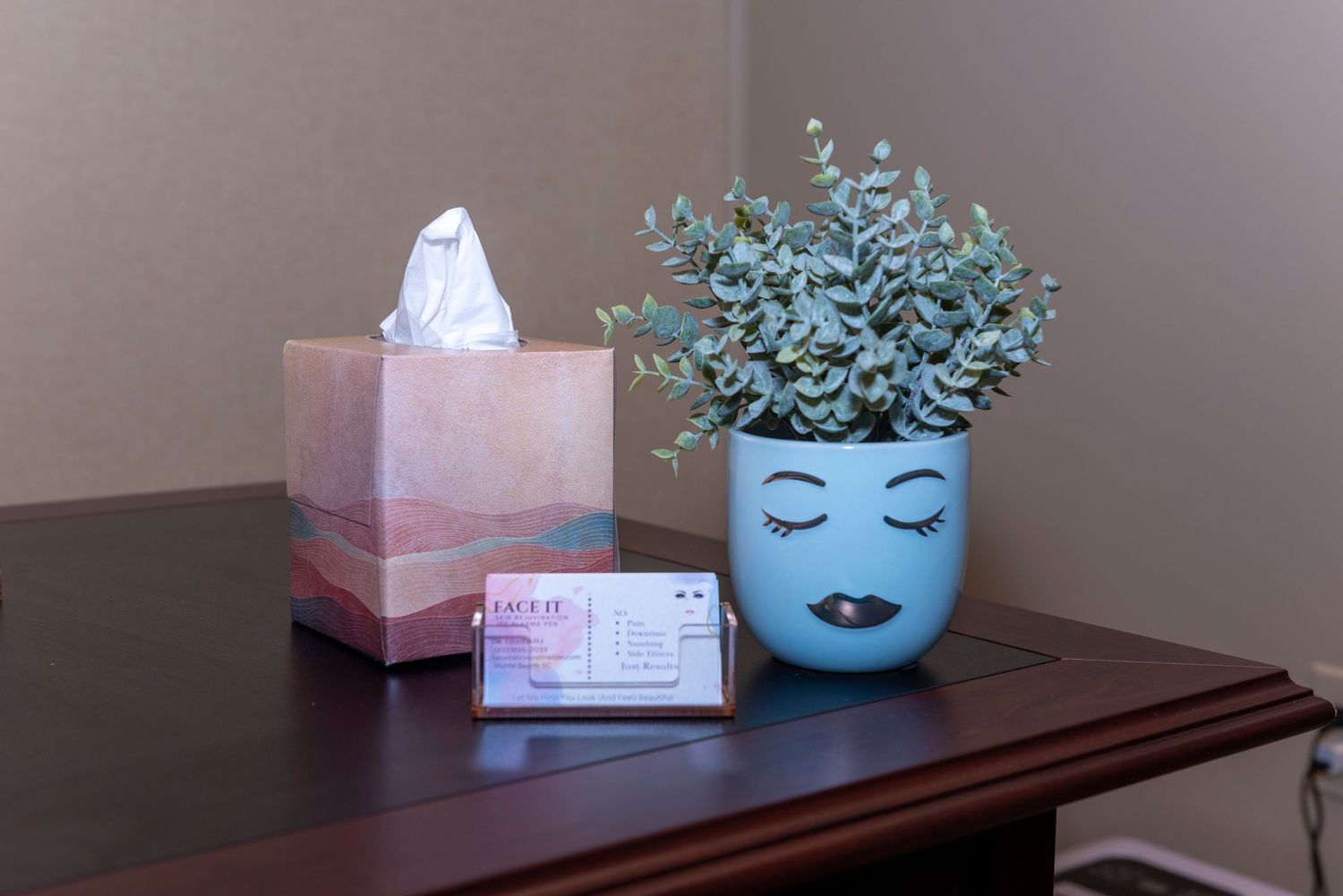 Teal decorative face planter with eucalyptus, pink tissue box, and business card holder on a wooden desk.