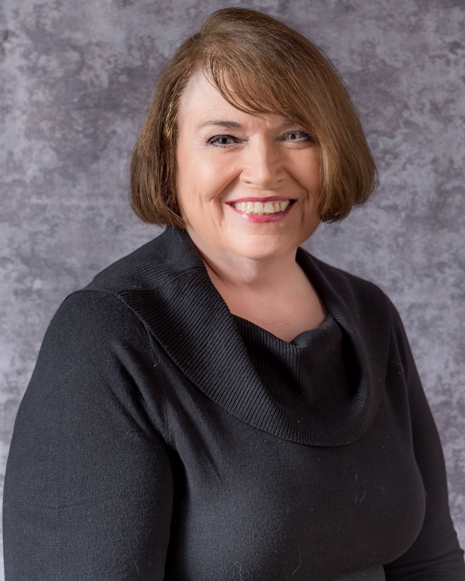 Professional headshot of a smiling woman with short brown hair wearing a dark cowl-neck sweater against a gray backdrop.