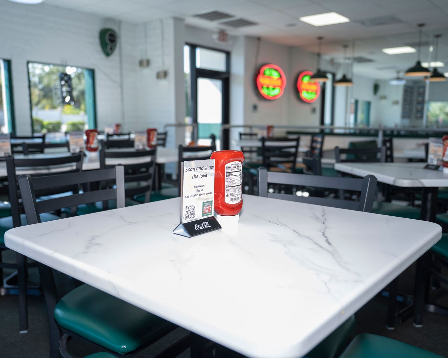 Bright restaurant dining room with marble tables, green chairs, and a red hot sauce bottle centerpiece.