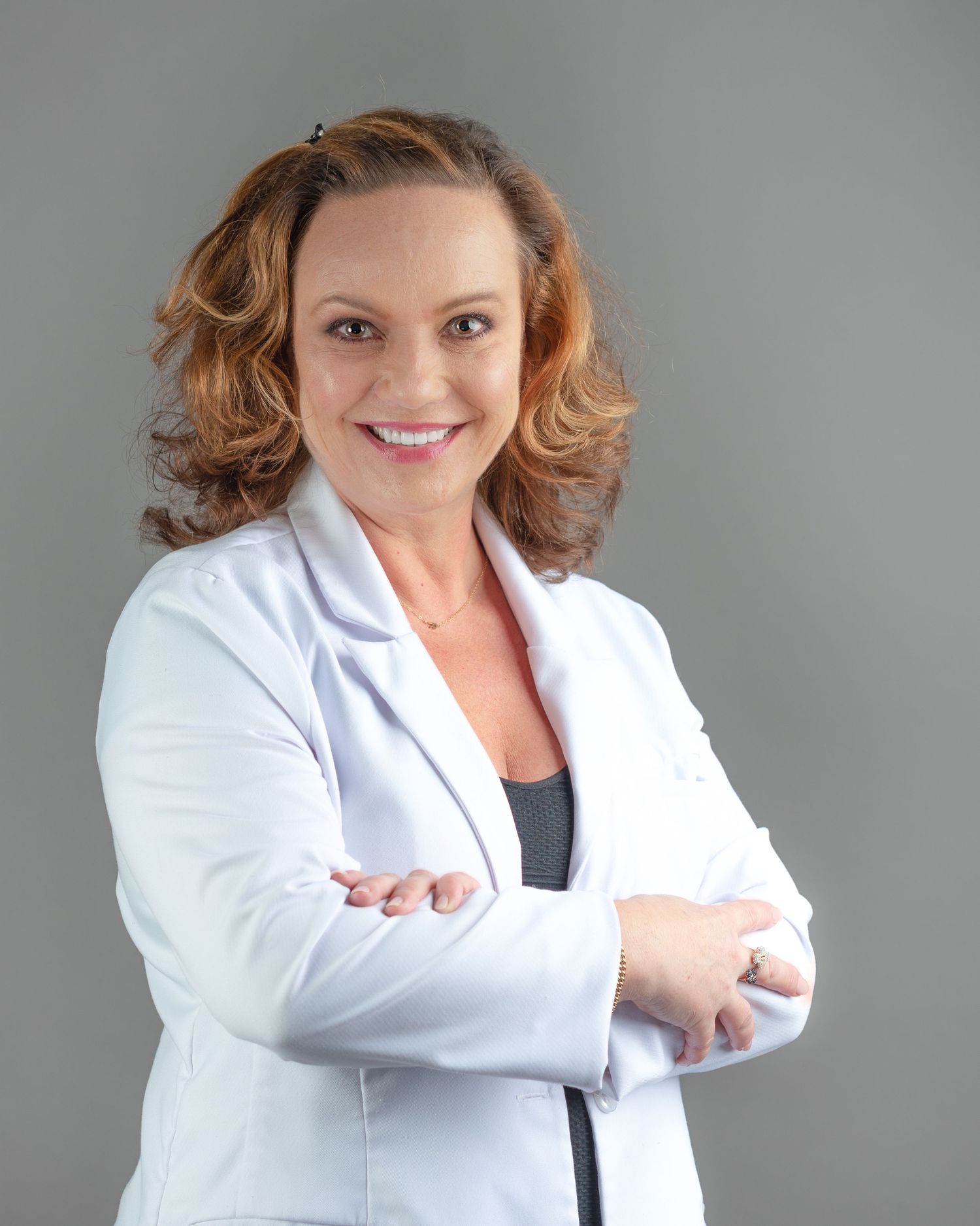 A smiling female doctor in a white lab coat stands with arms crossed against a gray background.