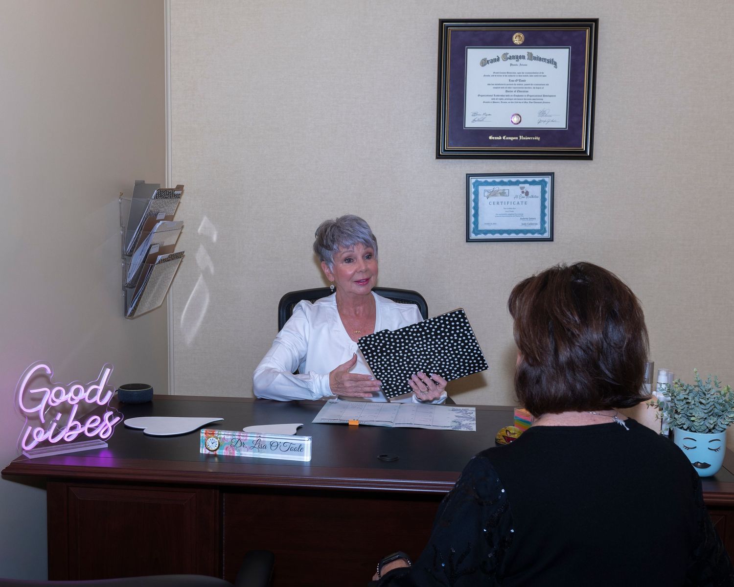Professional woman consultant at desk showing document to client in modern office with Good Vibes neon sign and diplomas.