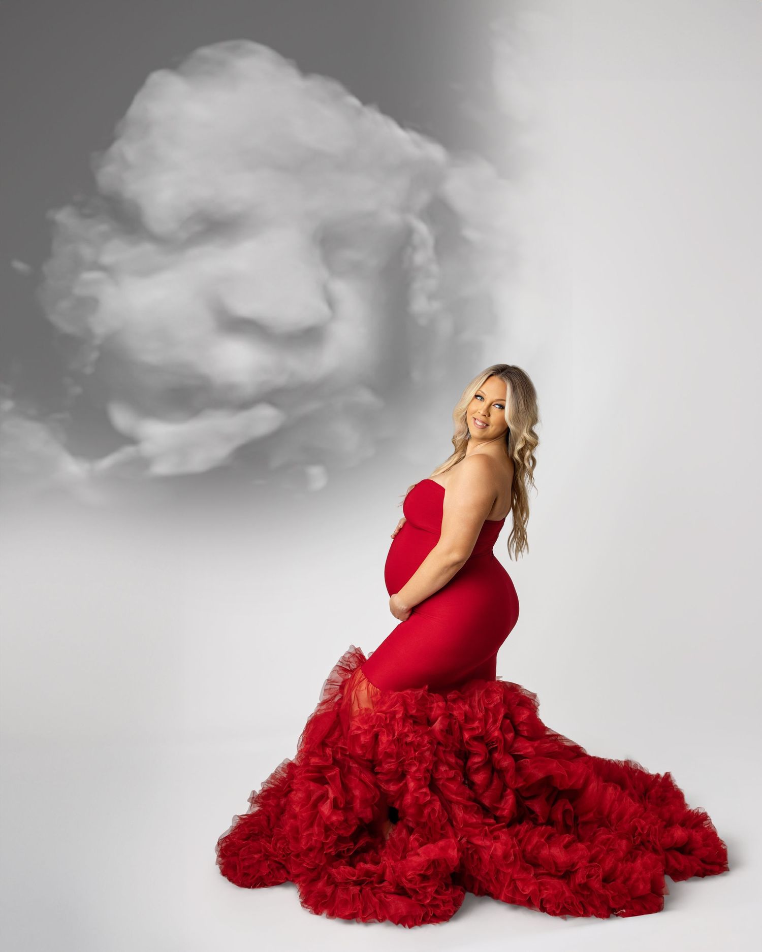 Pregnant woman in red ruffled gown poses gracefully with a cloud formation resembling a baby face in the background.