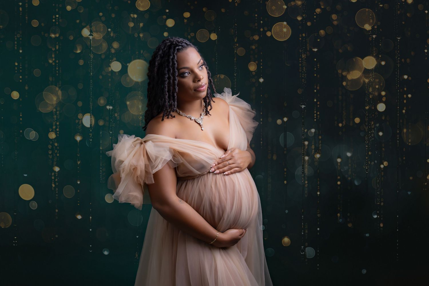 Pregnant woman in blush tulle gown cradling belly against dark teal backdrop with golden bokeh lights.