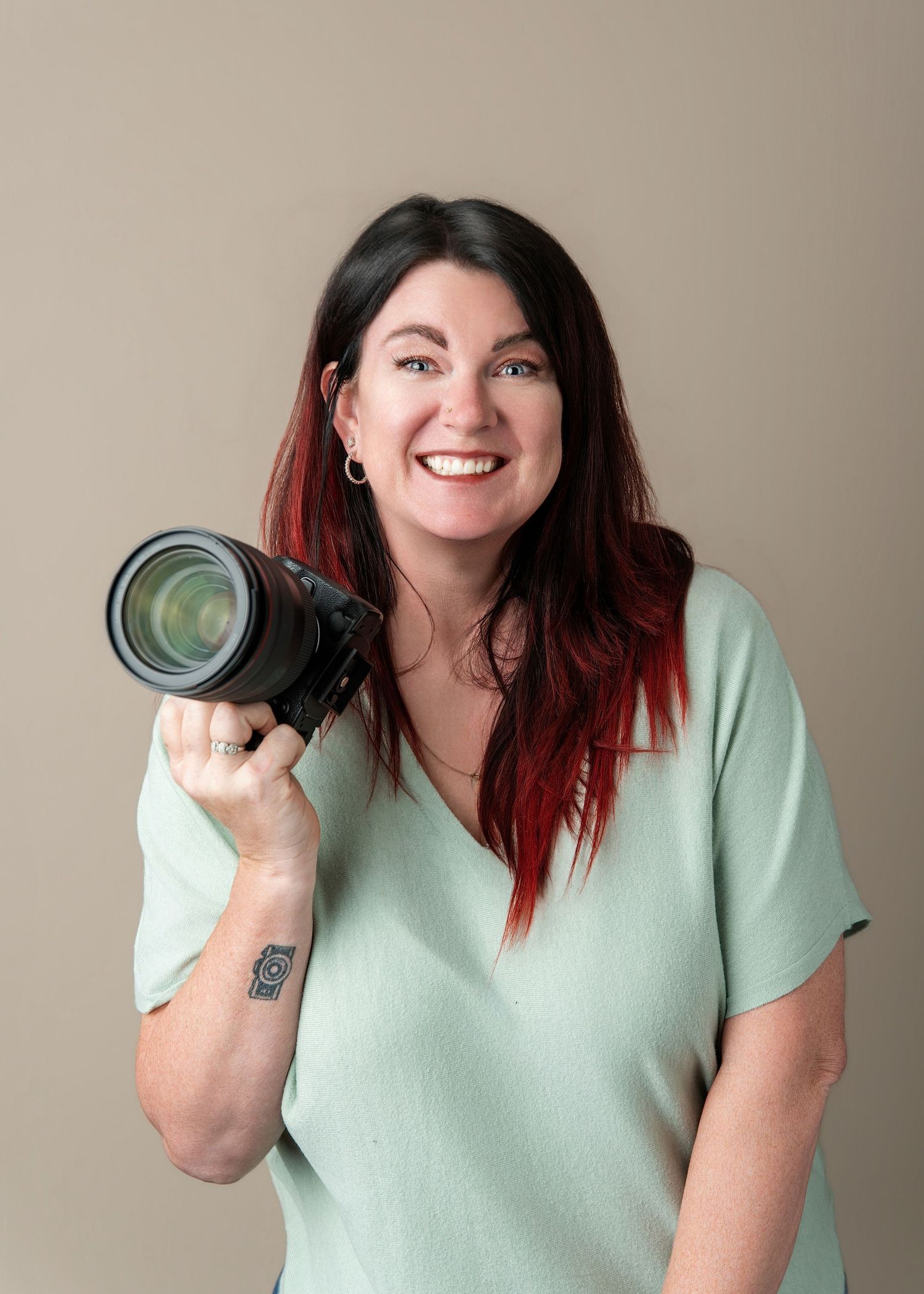 Smiling woman with red-tipped hair holds a professional camera, wearing a mint green top with a wrist tattoo.