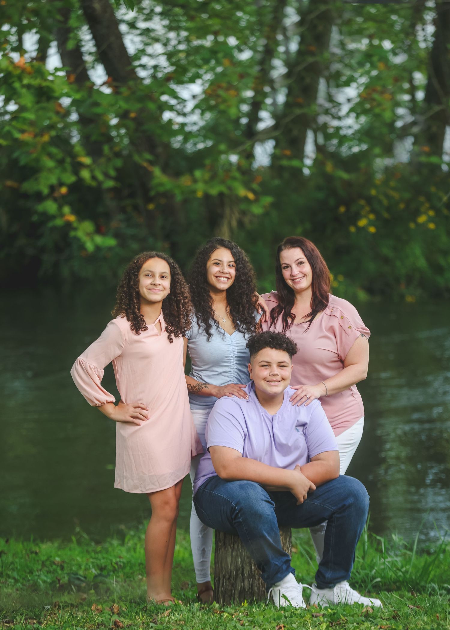 Four smiling siblings pose outdoors near a serene creek surrounded by lush green trees for a family portrait.