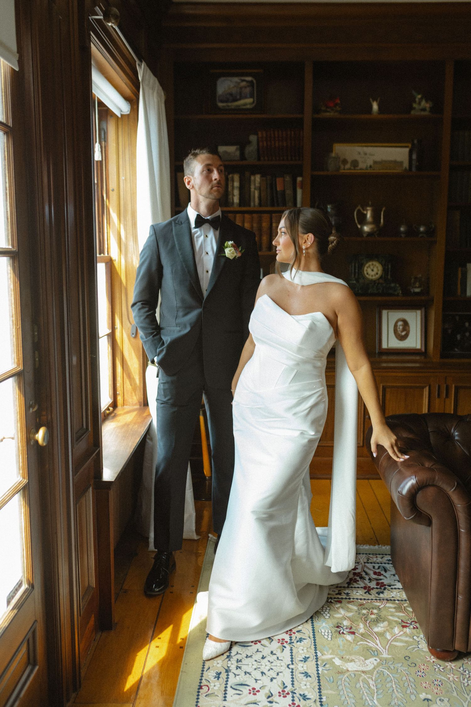A romantic wedding couple poses by sunlit windows in a vintage library room with ornate furniture and decor.