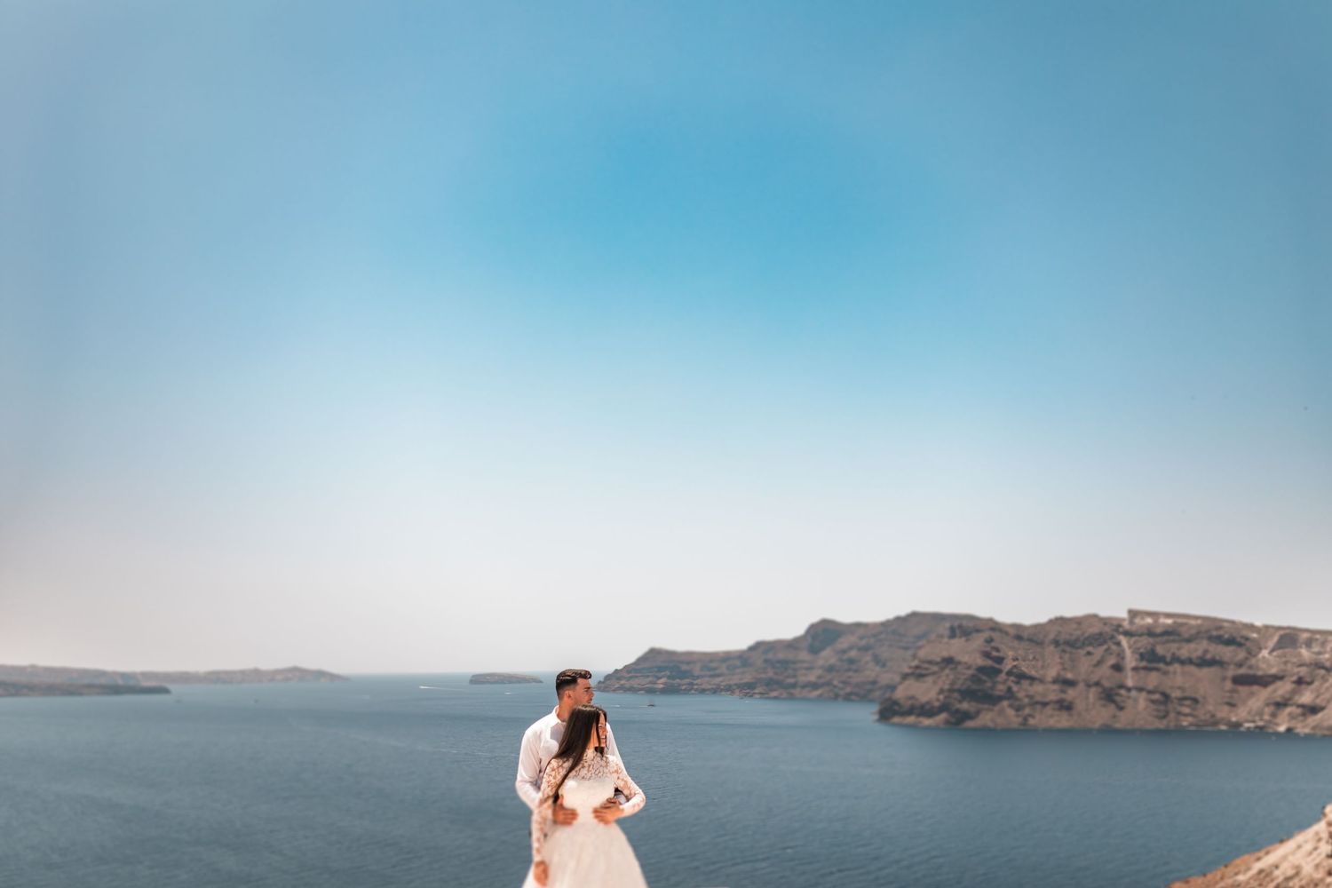 Couple in wedding attire standing on a cliffside overlooking the stunning blue waters of Santorini, Greece.
