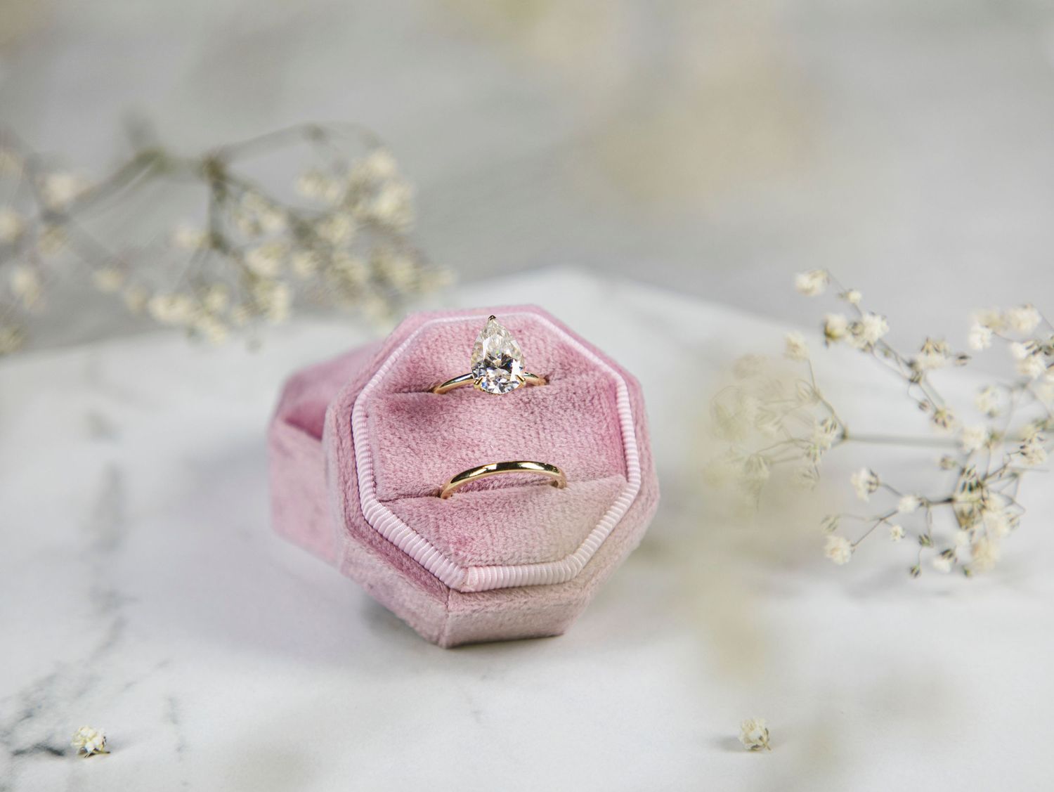 A pink velvet ring box containing a pear-shaped diamond engagement ring and a simple gold wedding band, with delicate wh
