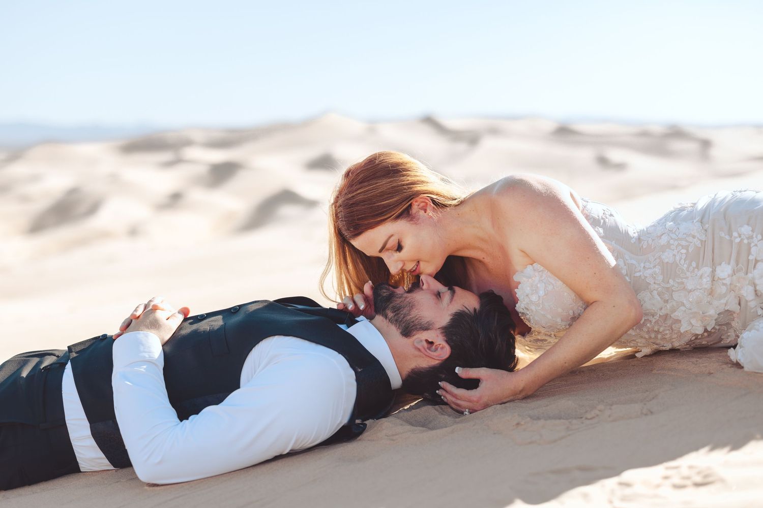 Couple in wedding attire lying romantically on sandy beach.
