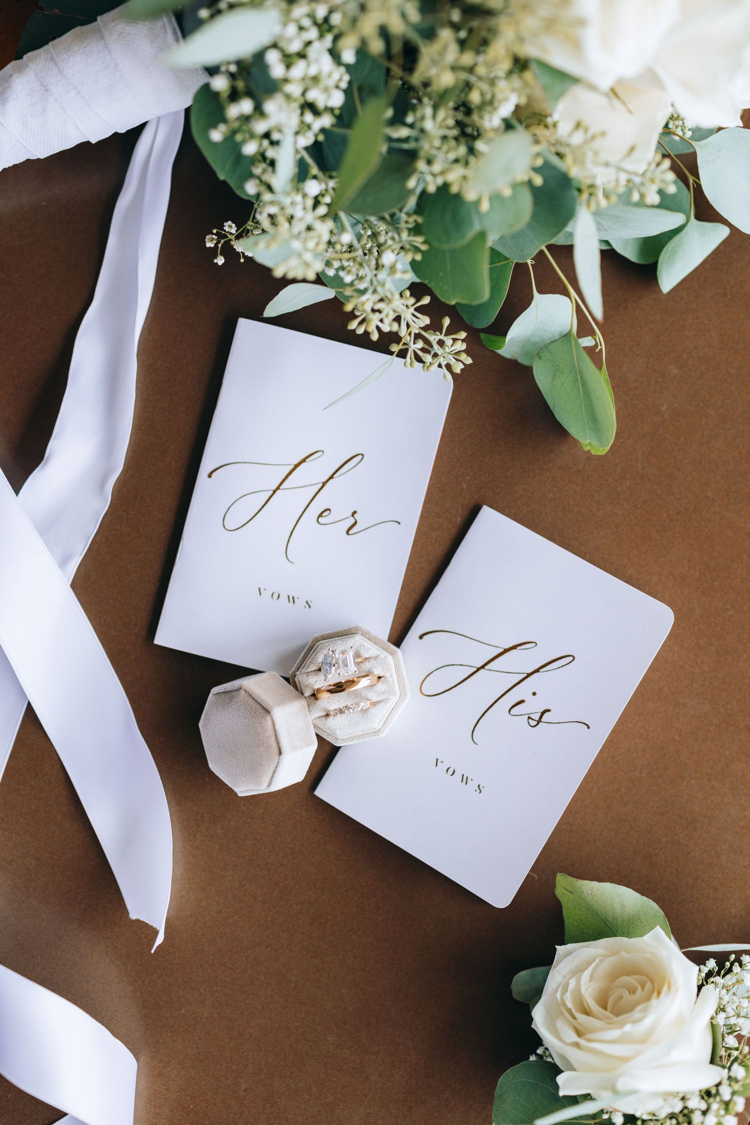 Wedding vow books labeled 'His' and 'Hers' styled with white flowers and ribbon.