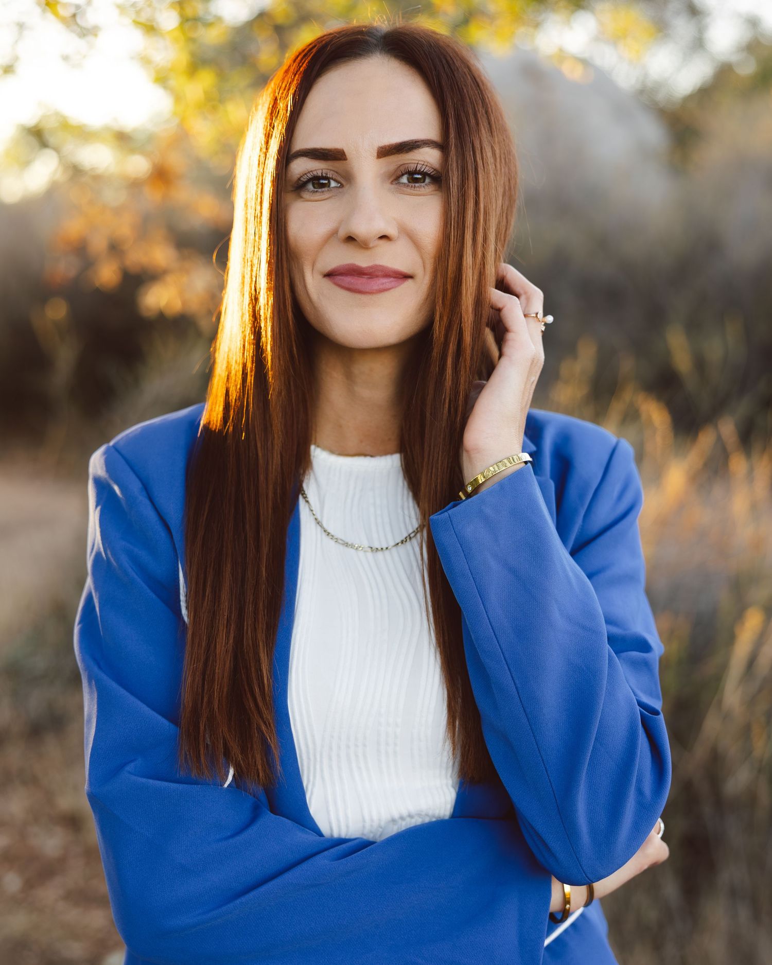 A woman in a bright blue blazer and white sweater poses outdoors during golden hour.