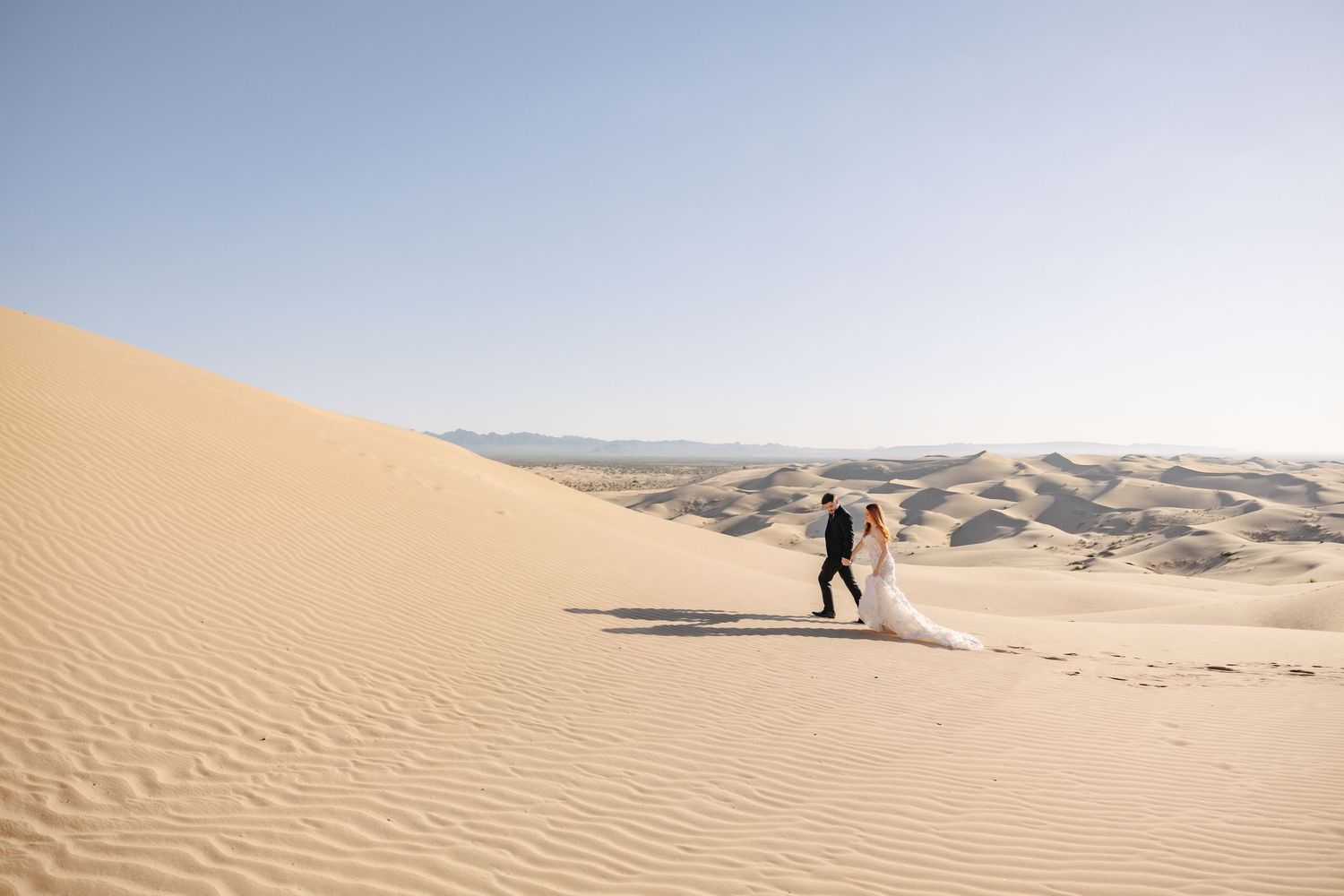 A couple walking through sweeping desert sand dunes under a bright sky.