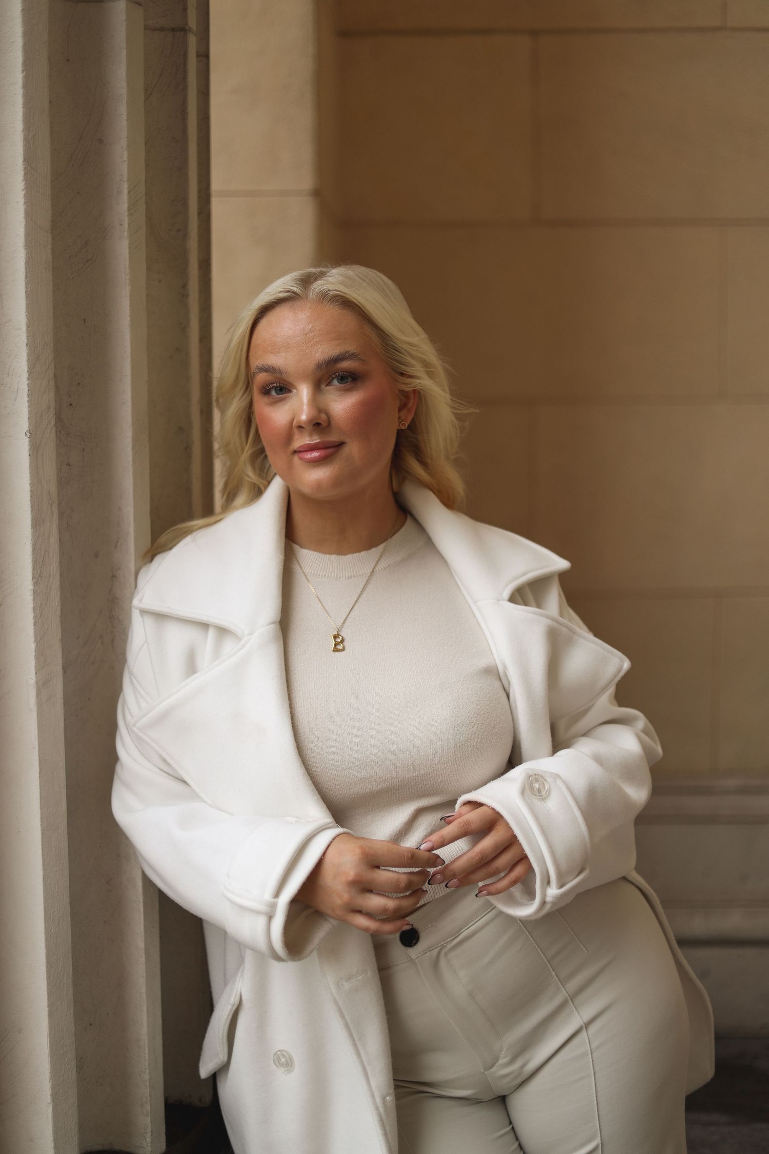 Fashionable woman in white winter outfit poses elegantly against stone column backdrop.