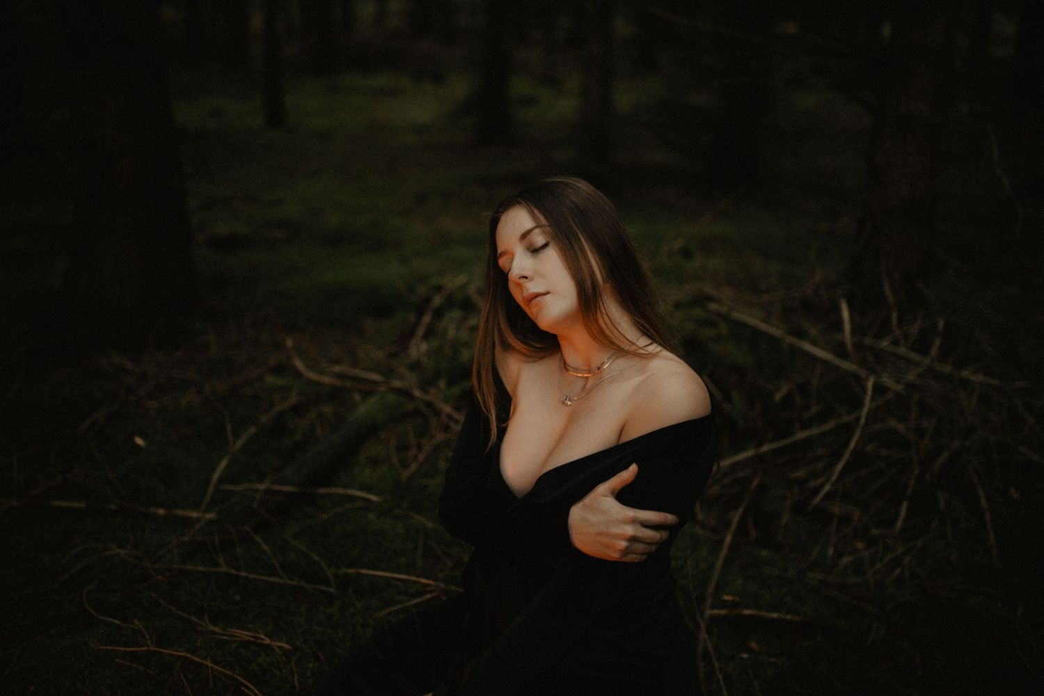 Moody portrait of woman in black dress against dark forest background.