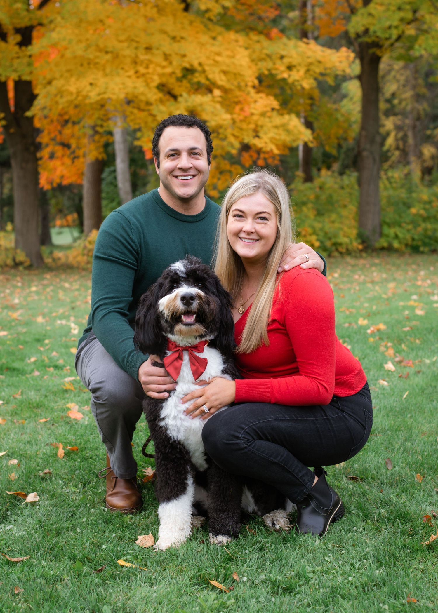 A black and white dog wearing a red bowtie poses with owners during autumn outdoor photo session.